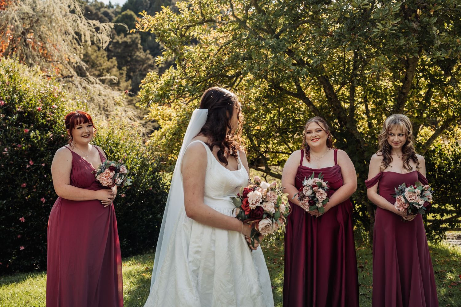 Bride in white gown with bridesmaids in burgundy dresses holding bouquets outdoors at a garden wedding.