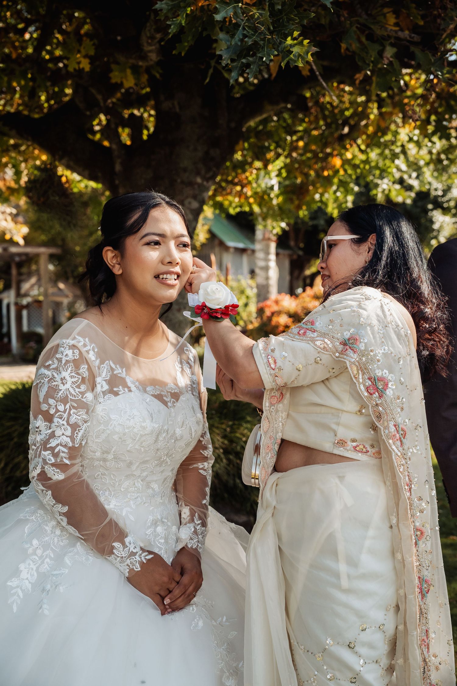 Smiling bride in lace wedding dress being touched up outdoors by woman in white saree.