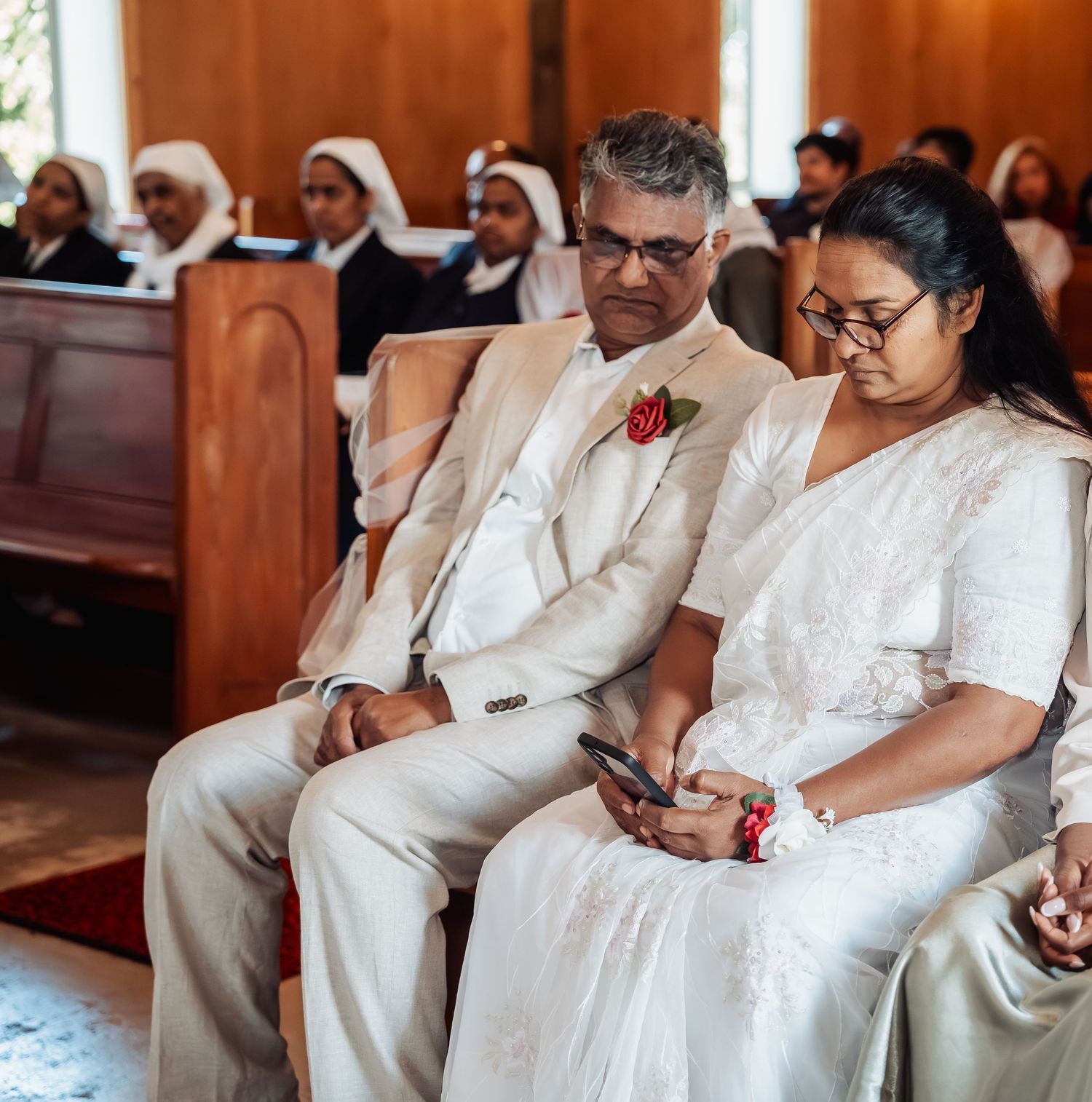Elegant couple in white attire seated in church pew during wedding ceremony with nuns visible in background.