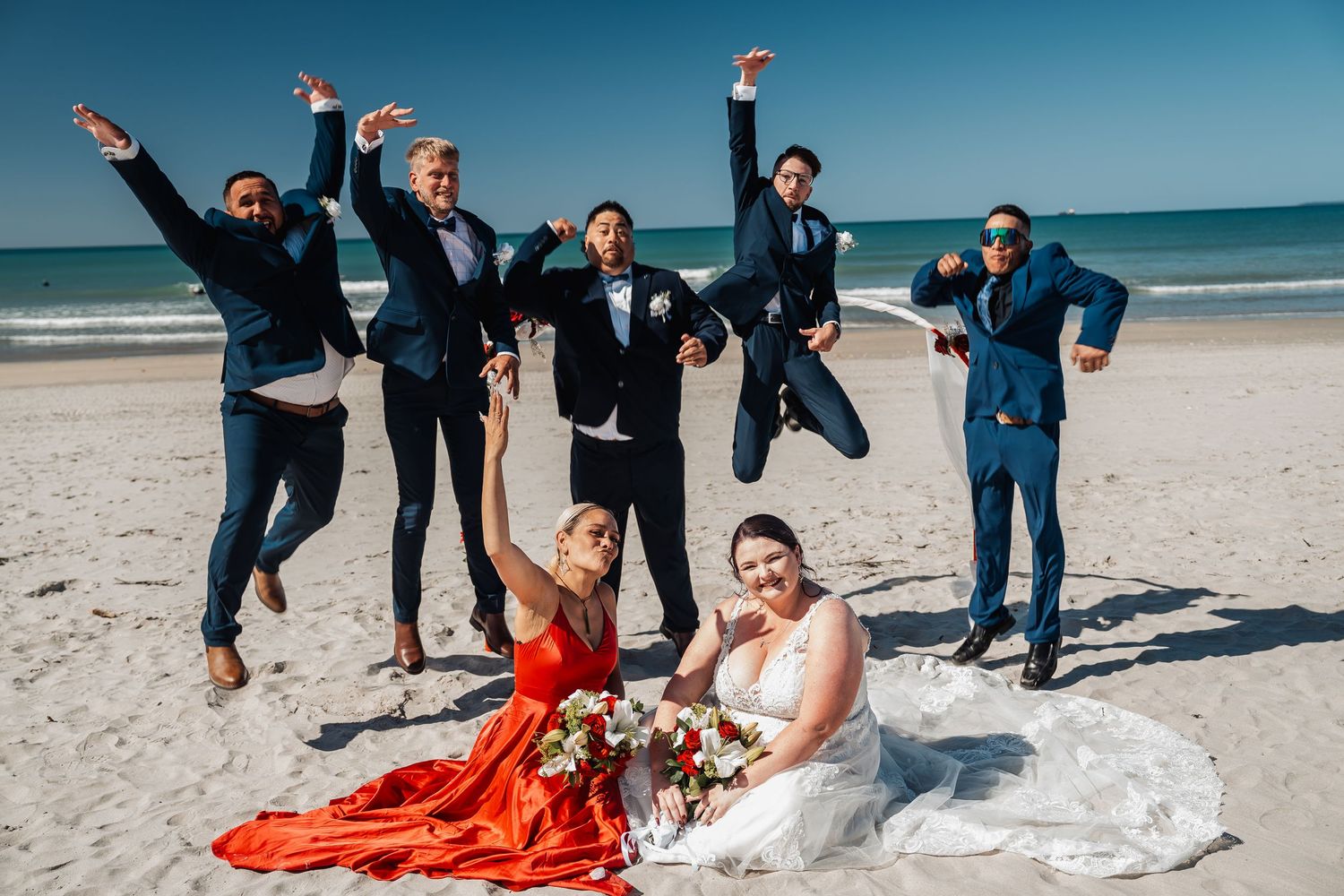 Wedding party celebrates joyfully on white sandy beach with ocean backdrop.