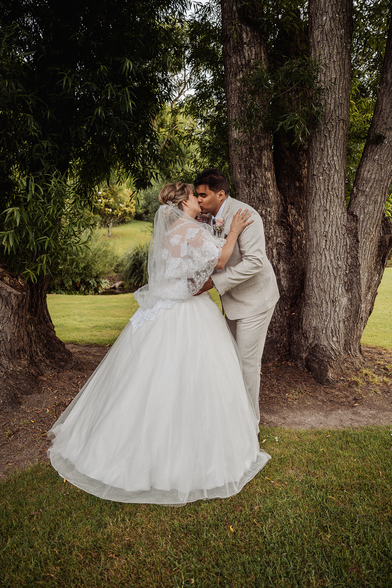 Romantic wedding moment captured under tall trees in outdoor garden setting.