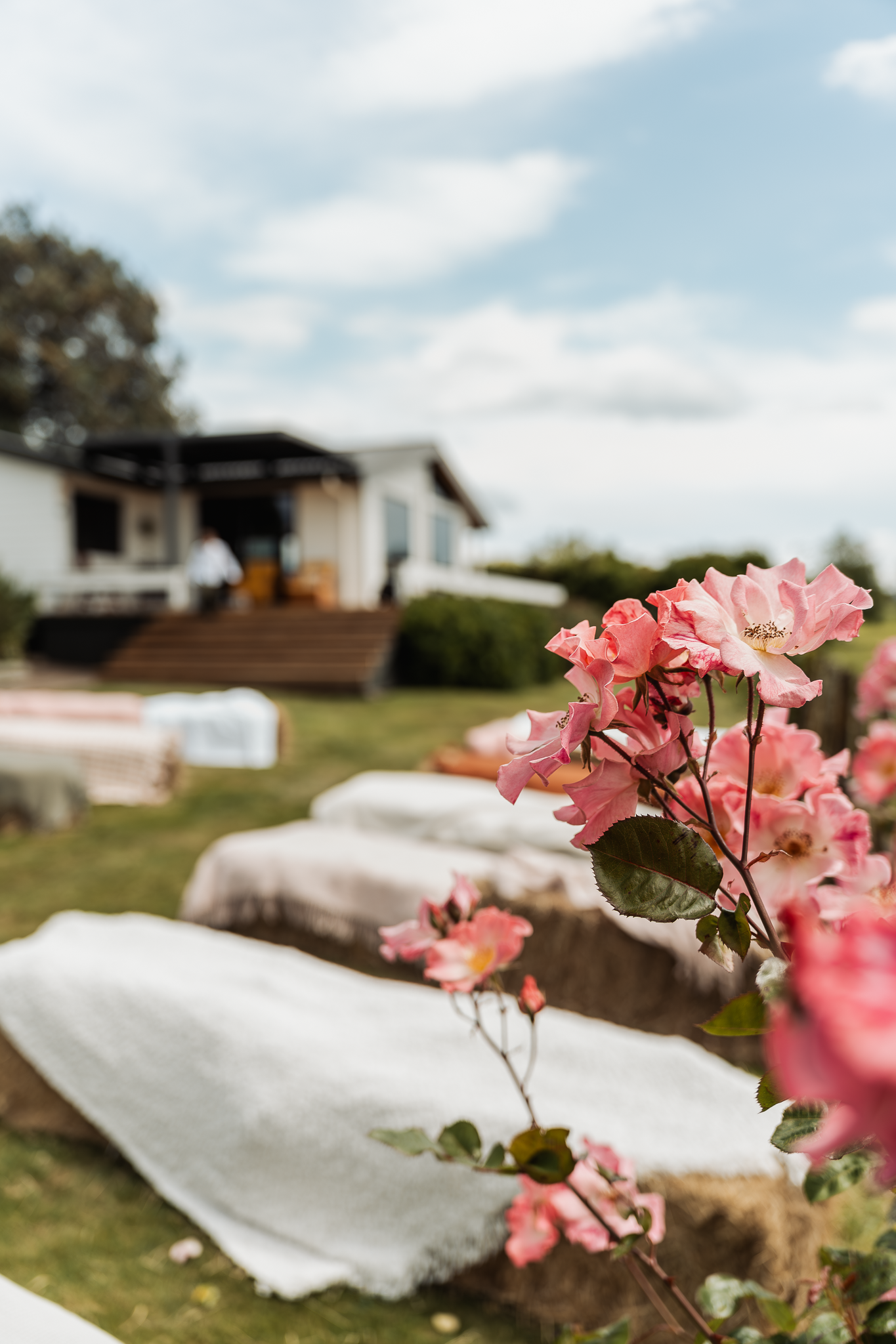 Pink flowers bloom in front of a rustic outdoor wedding venue with white chairs arranged on the lawn.