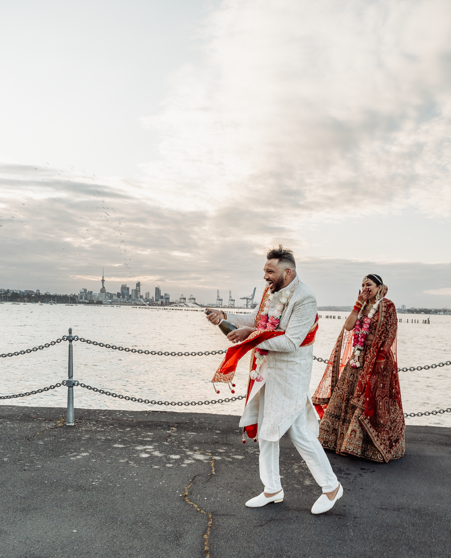 Couple in traditional Indian wedding attire enjoying a moment on waterfront deck with cityscape background.
