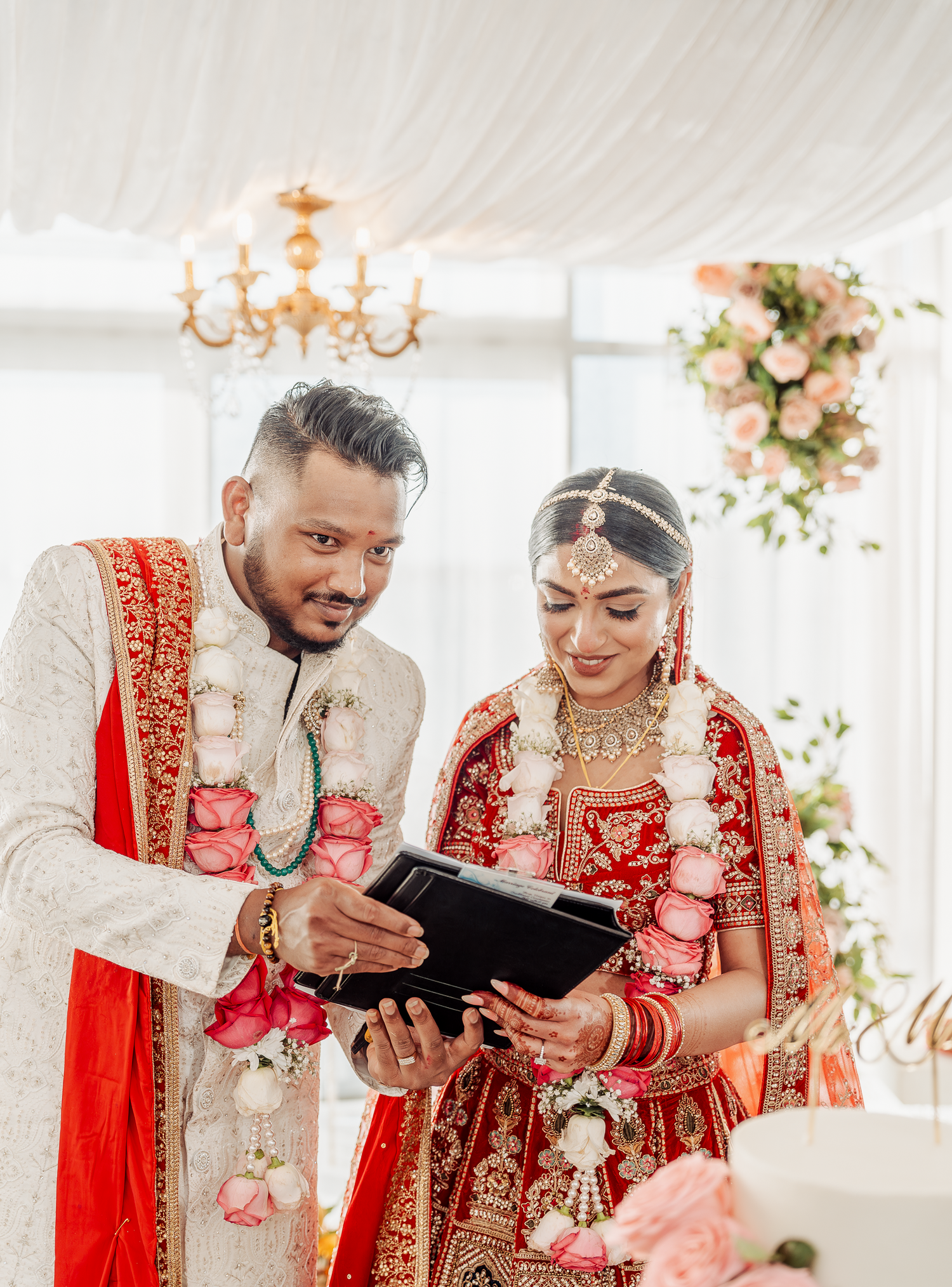 Couple in traditional wedding attire reviewing paperwork during their ceremony under chandelier.