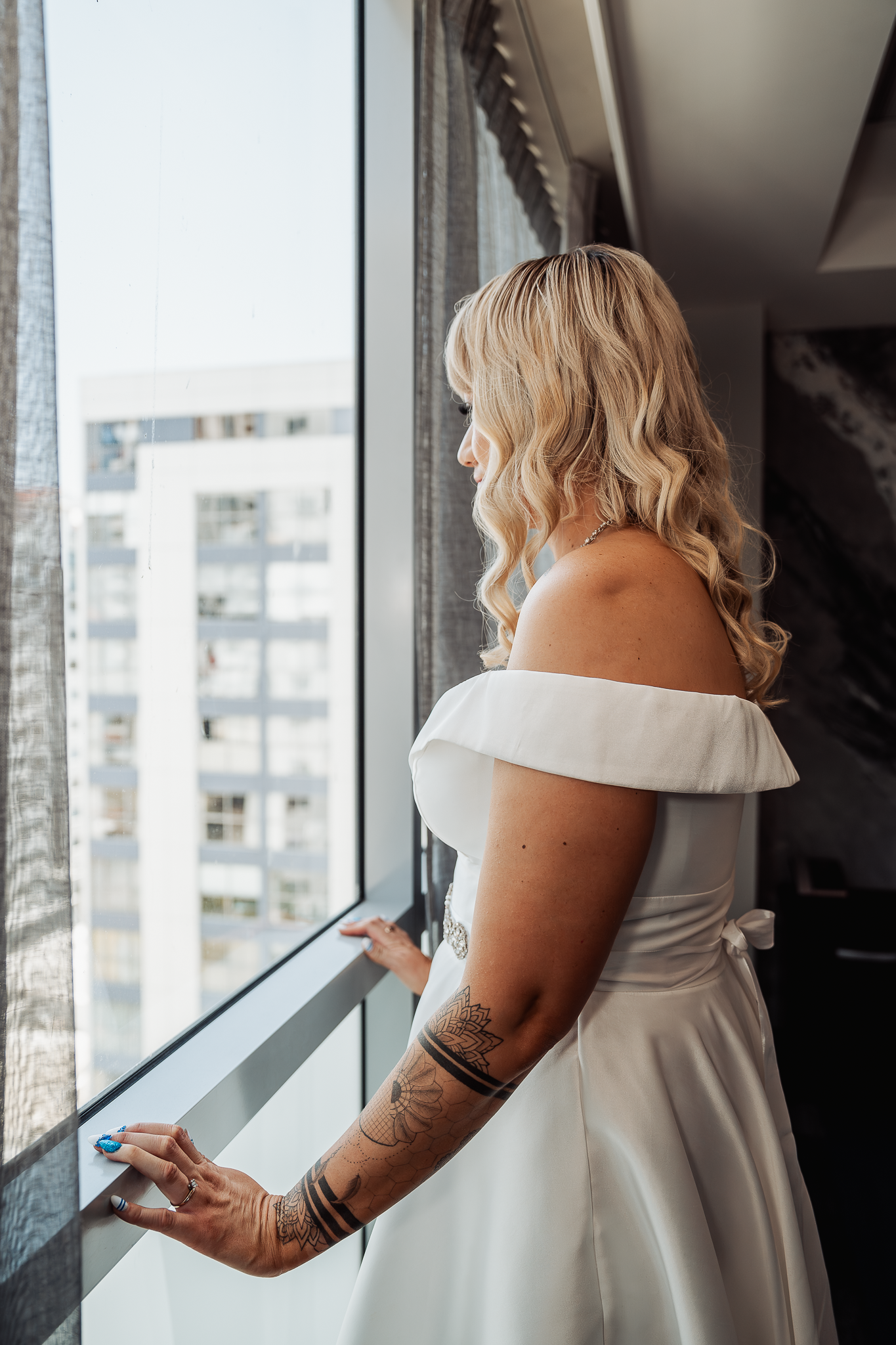 Bride in white off-shoulder dress gazes out high-rise window.