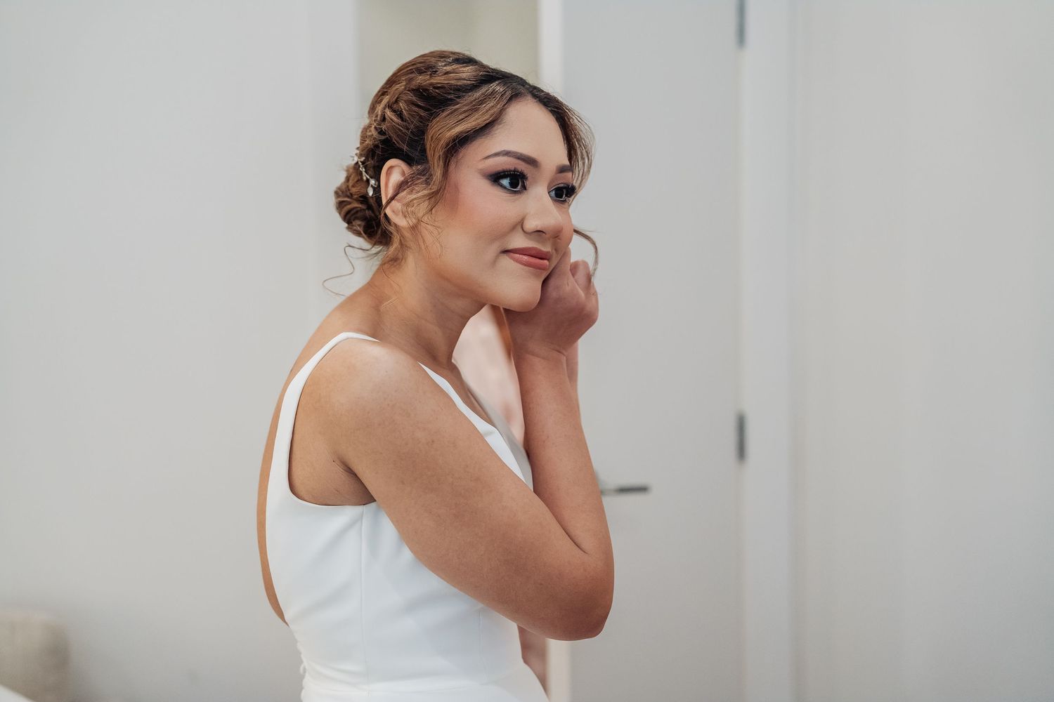 Someone in a white sleeveless dress touches their face while smiling during what appears to be a bridal preparation moment.