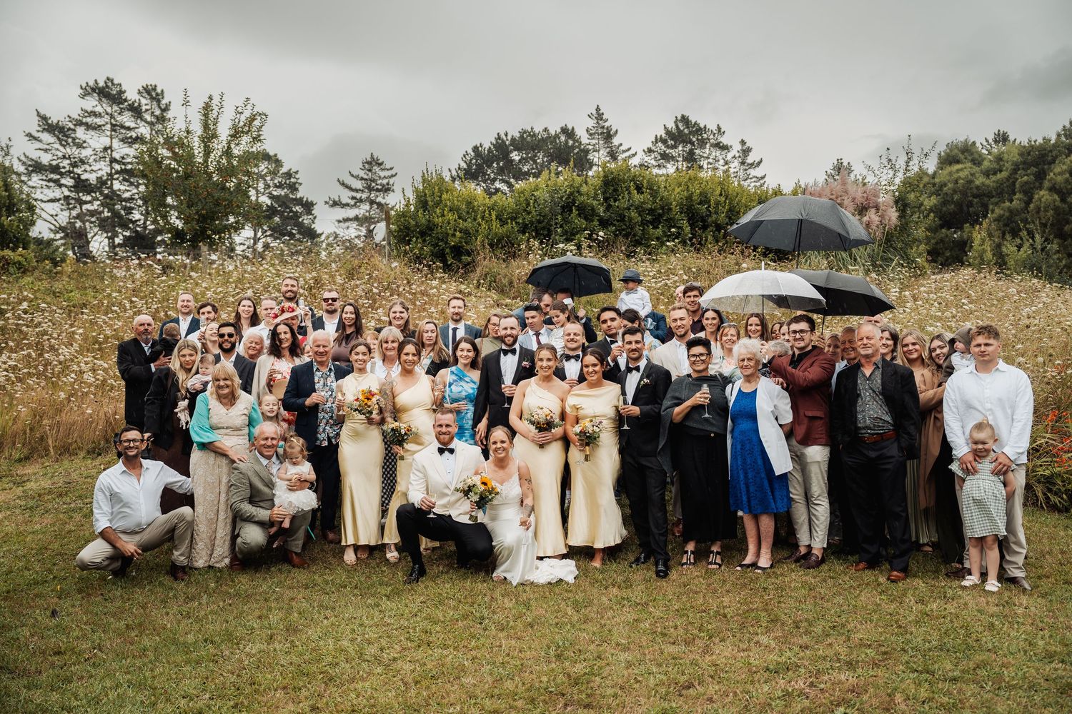 Large wedding group photo outdoors in a field with guests holding umbrellas on a cloudy day.