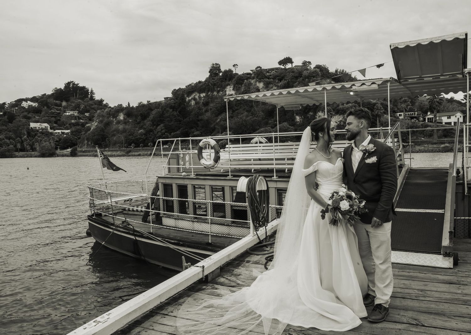 Bride and groom sharing romantic moment on dock beside vintage boat in black and white.