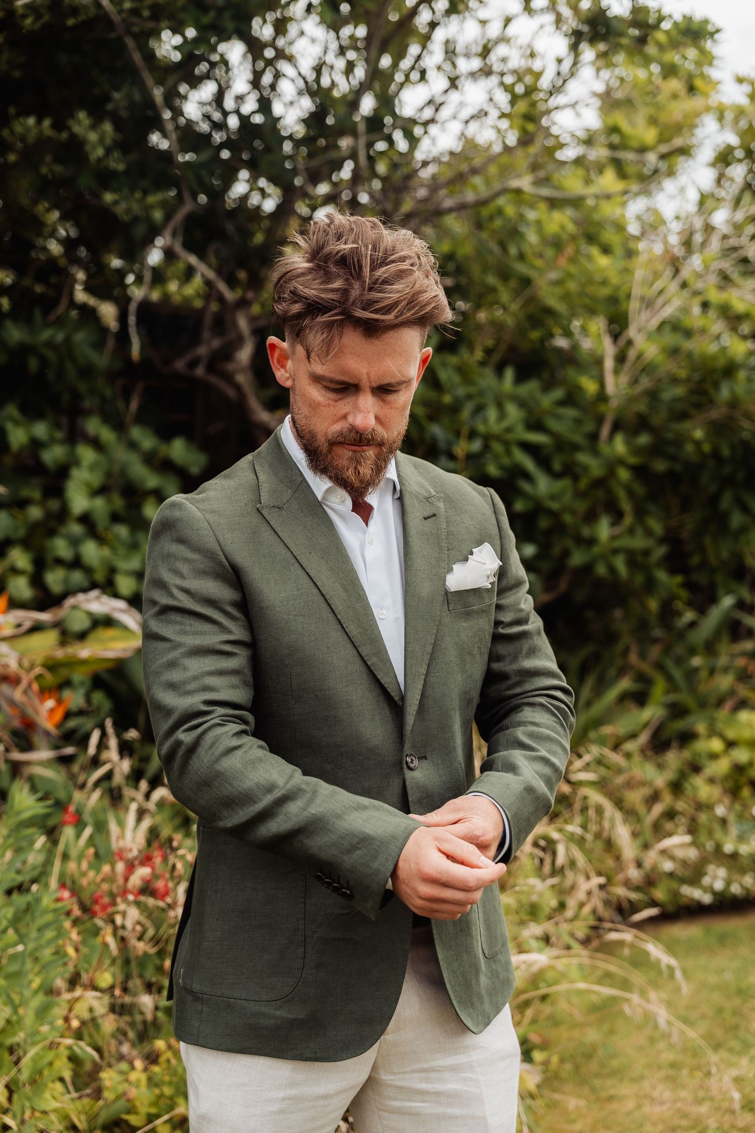 Bearded groom in green blazer adjusts his sleeve in a lush garden setting.