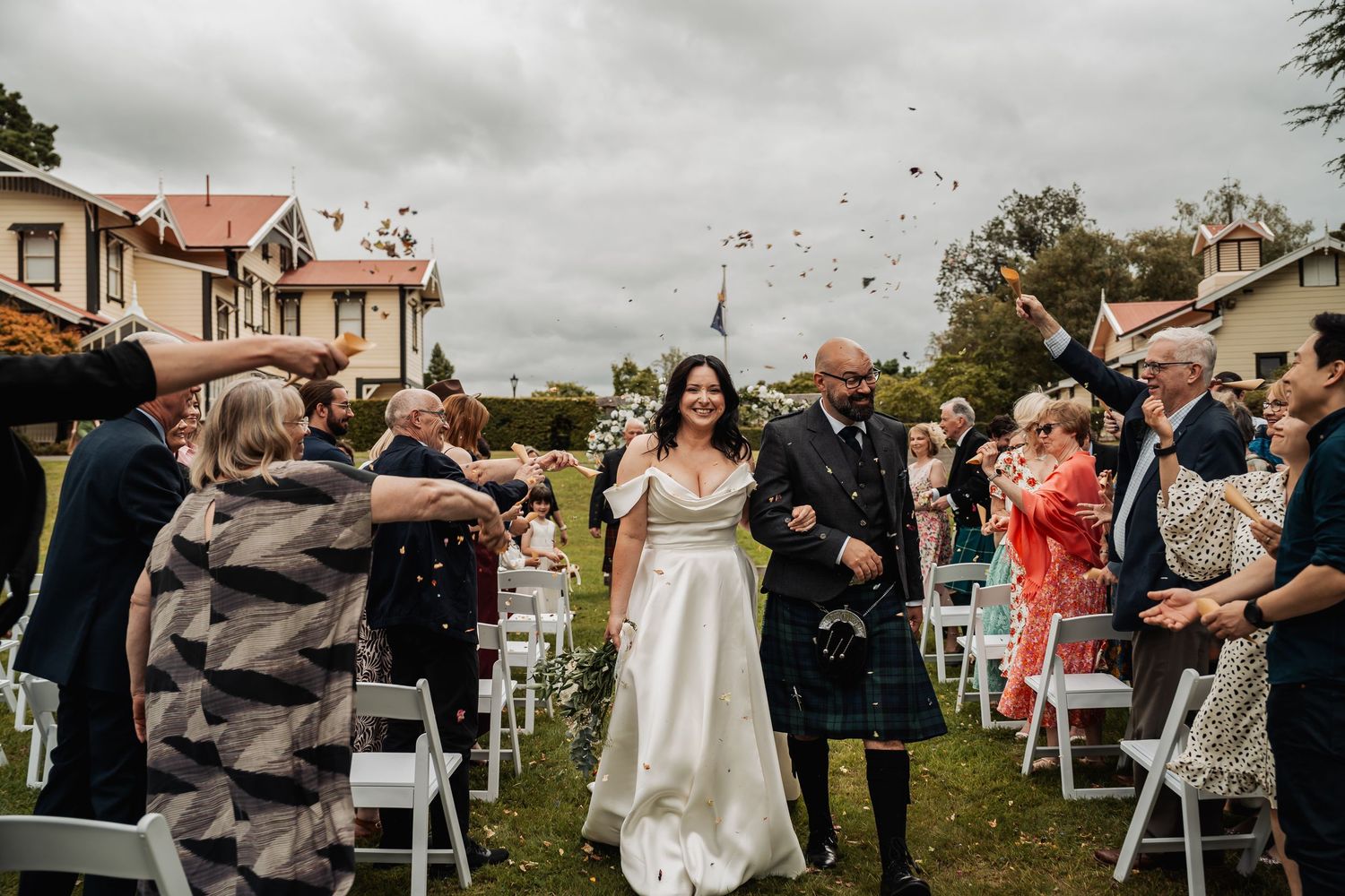 Bride and groom walk through confetti shower at outdoor wedding ceremony on cloudy day.