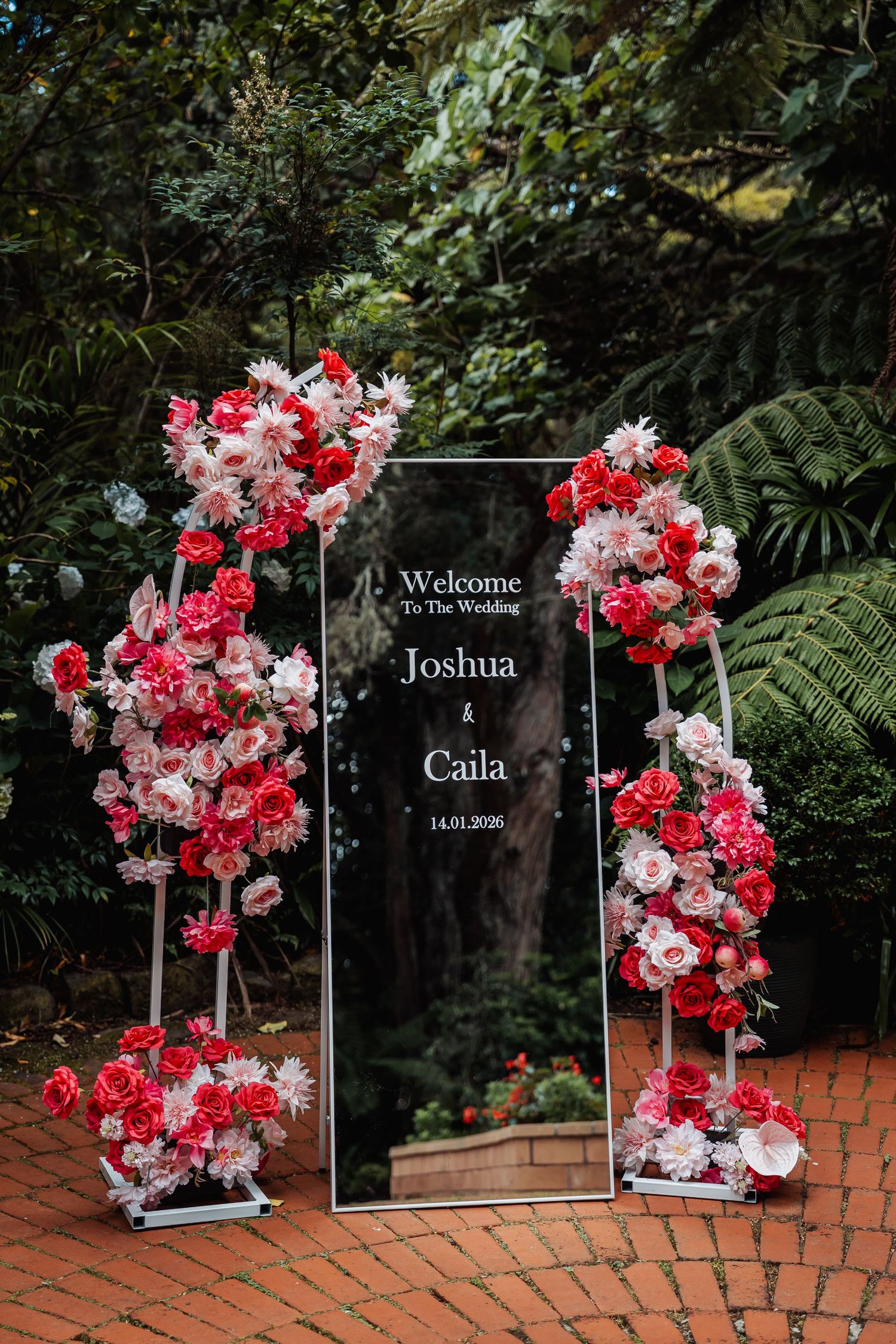 Elegant wedding welcome sign for Joshua and Caila adorned with pink and red floral arrangements on a mirrored display outdoors.