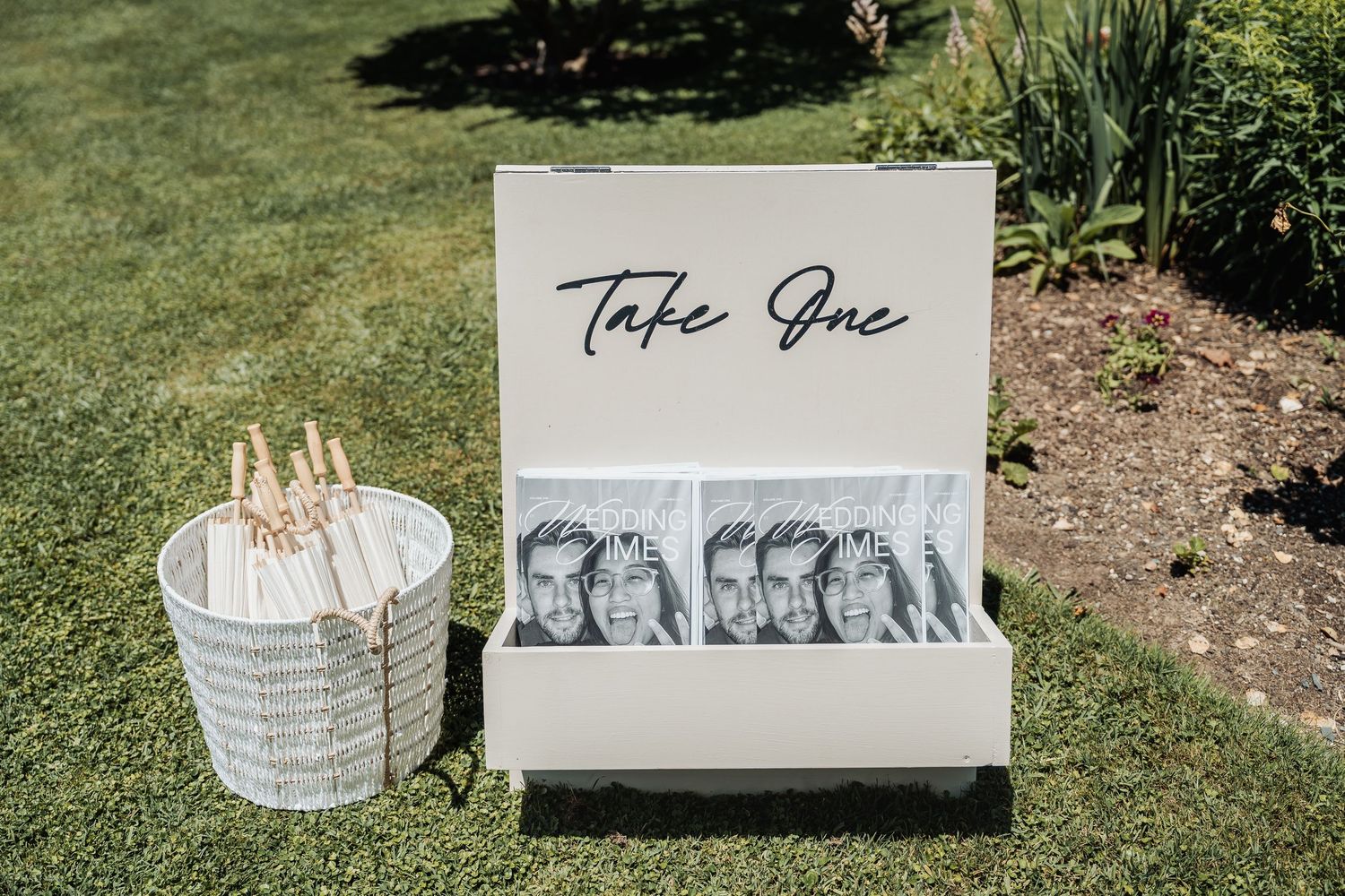 A white wedding program display box labeled Take One sits on grass beside a wicker basket with fan programs.