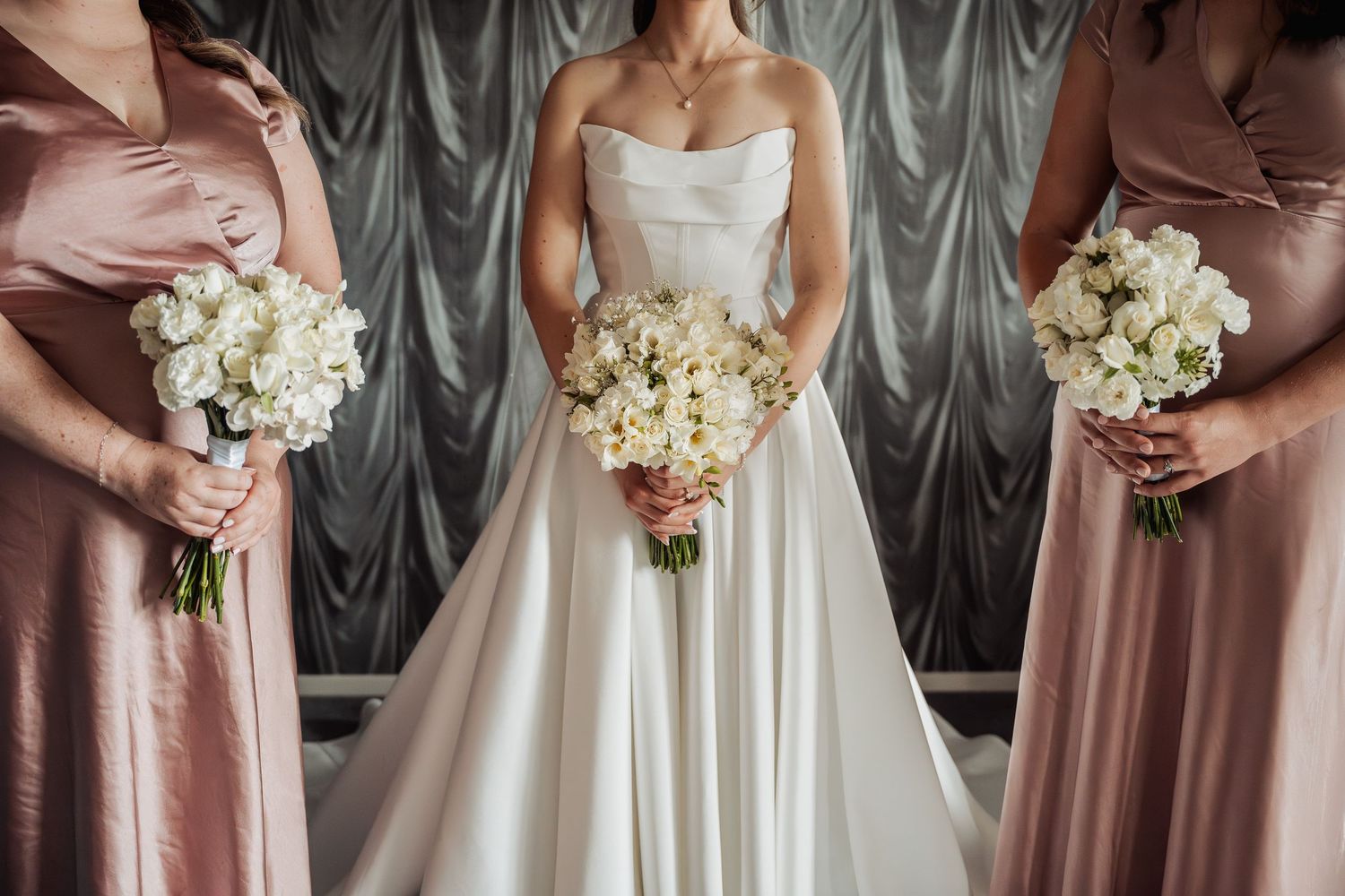 Bride in a strapless white gown flanked by two bridesmaids in blush pink dresses holding white bouquets.