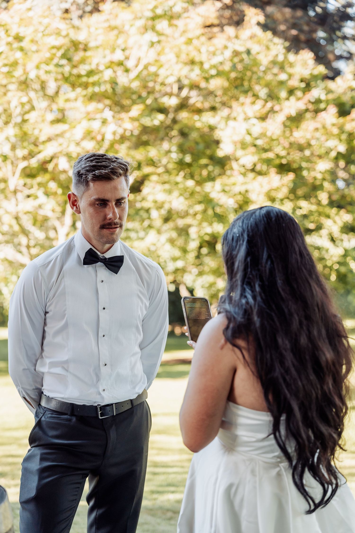 Groom in white shirt and bow tie listens attentively as bride reads wedding vows outdoors.