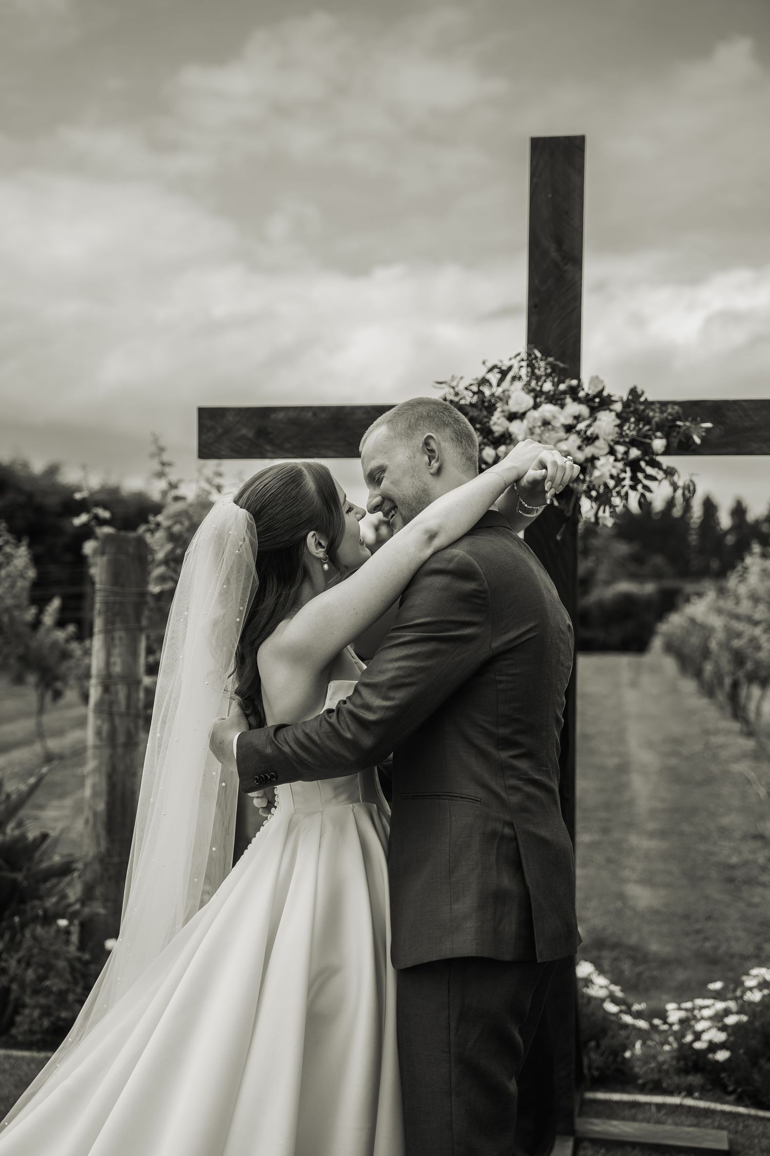 Black and white photo of bride and groom embracing before a wooden cross adorned with floral arrangements.