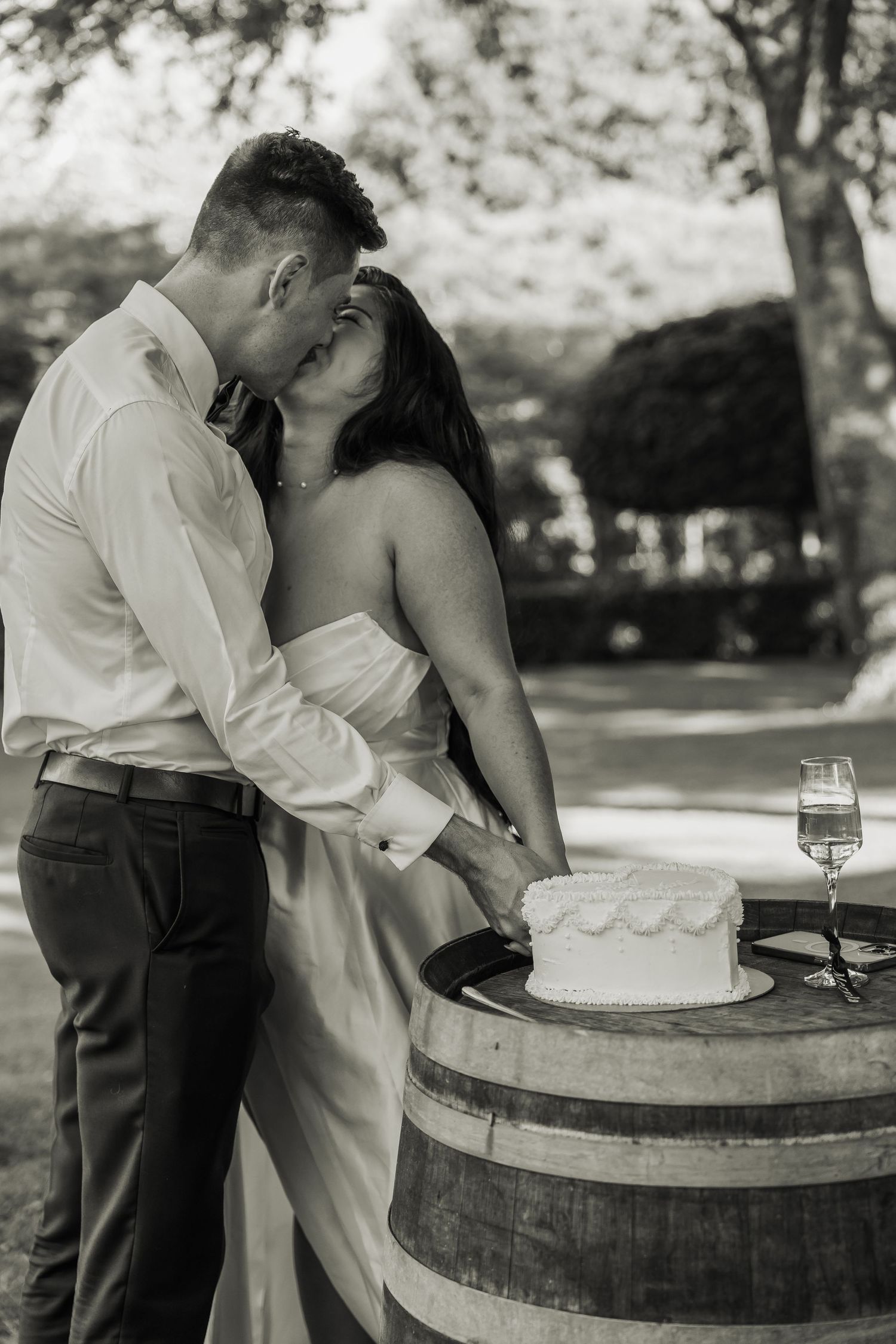 Black and white photo of bride and groom cutting wedding cake on a rustic barrel outdoors.