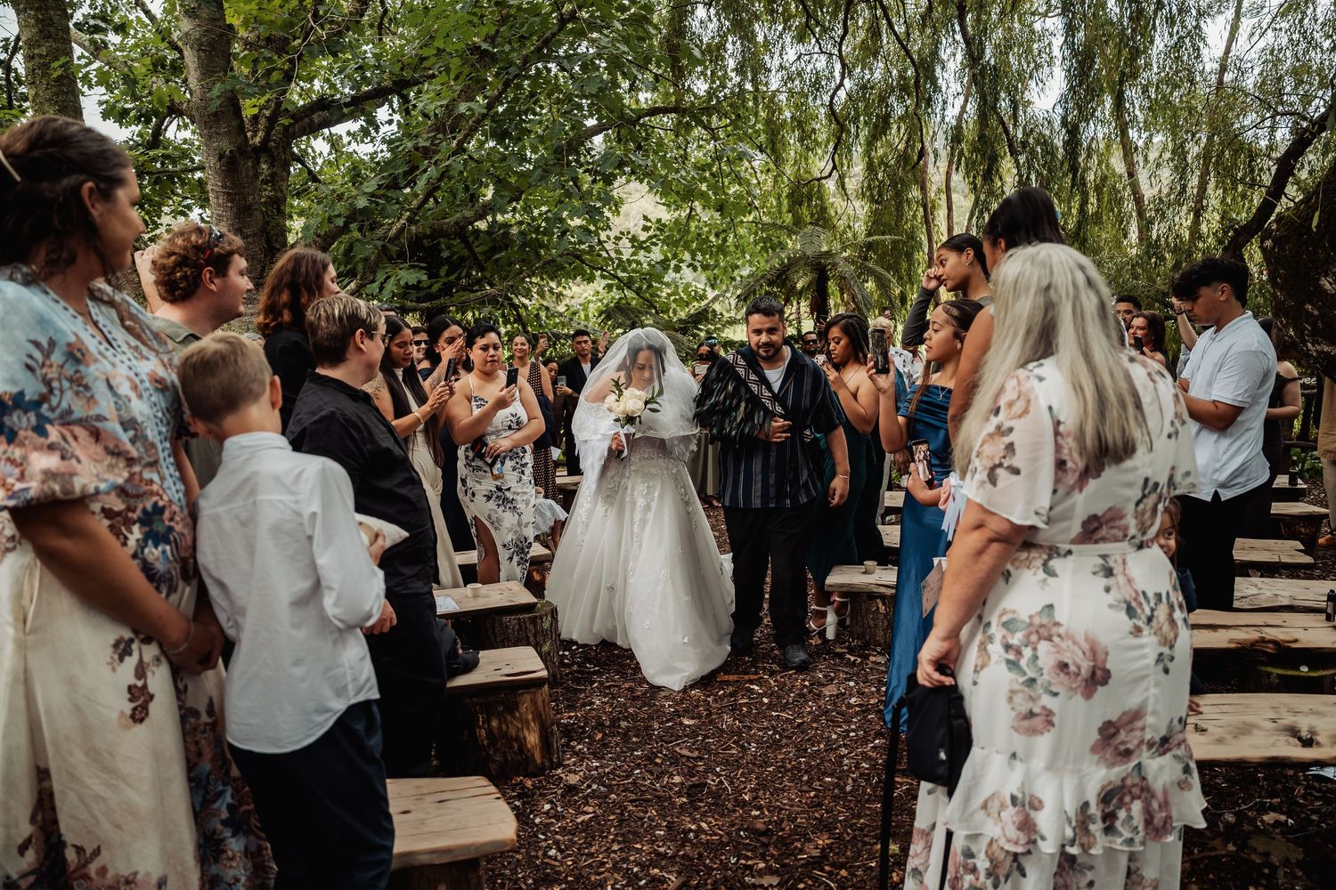 Guests gather outdoors for a woodland wedding ceremony as the bride walks down the aisle.