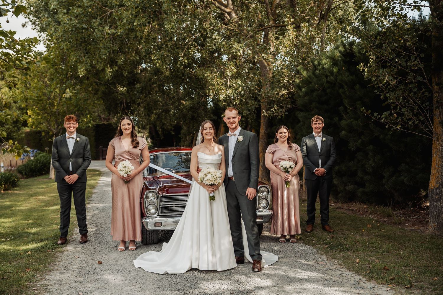 Wedding party poses outdoors with bride and groom centered, bridesmaids in blush gowns beside vintage red car.