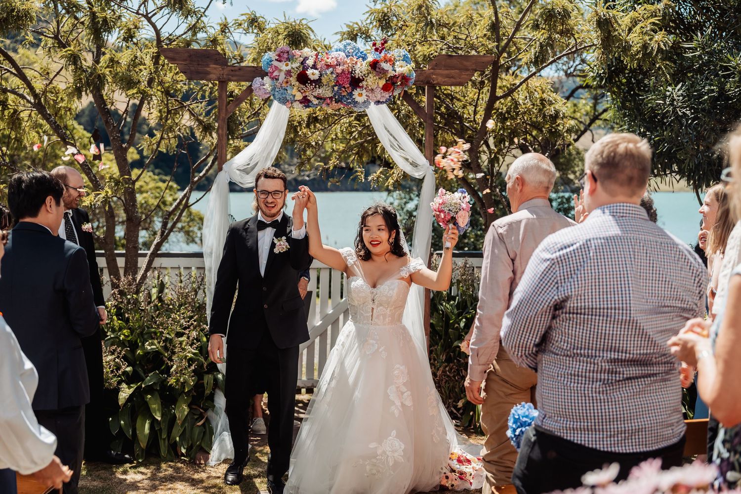 Newlyweds raising hands in celebration as they walk back down aisle under floral arch by lake.