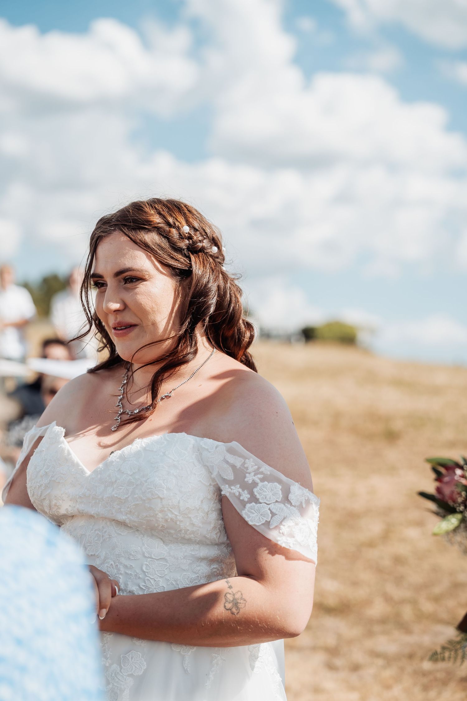 Bride in off-shoulder lace wedding dress with hair accessories at outdoor ceremony under blue sky.