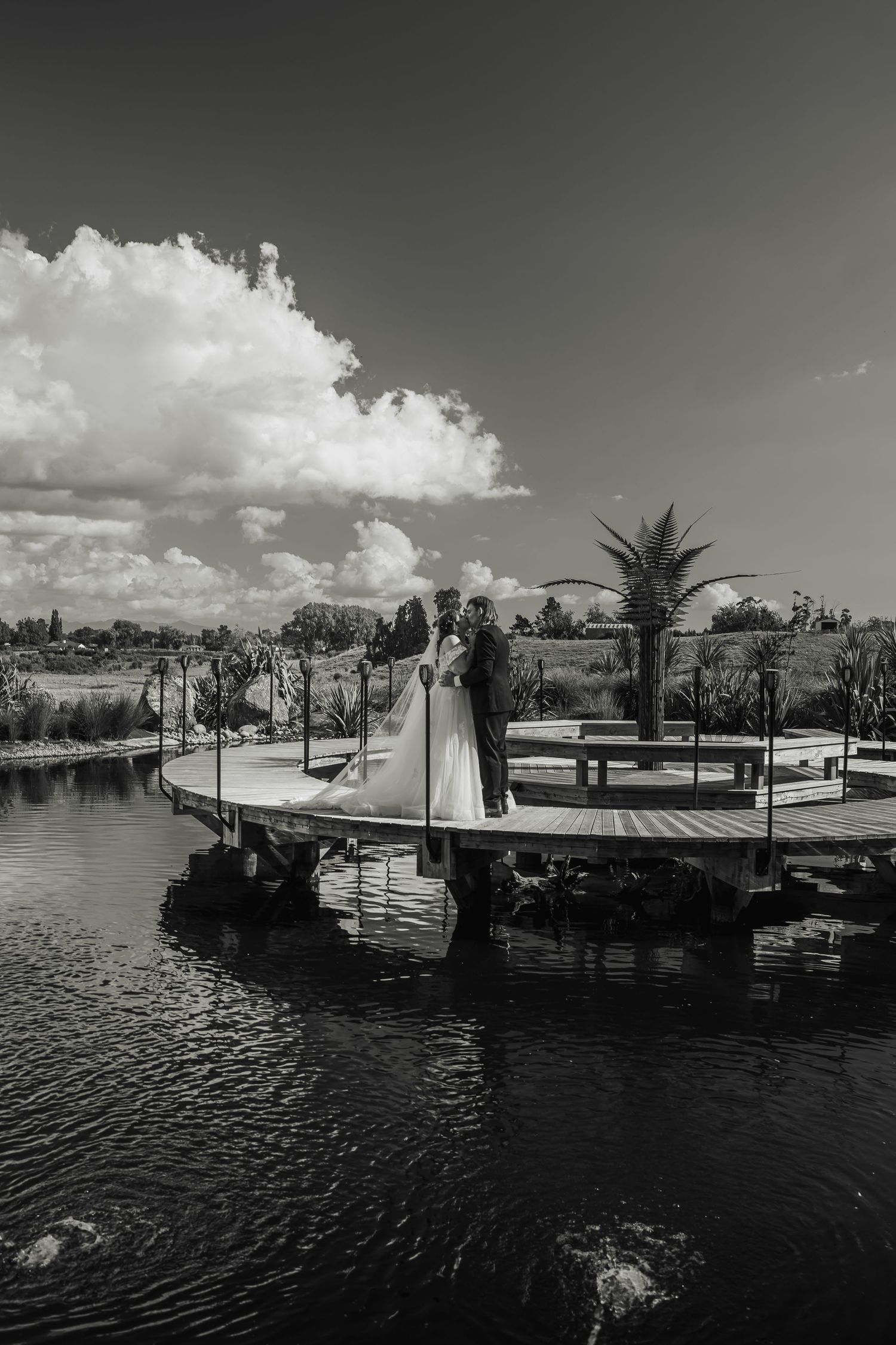 Bride and groom embrace on waterfront dock surrounded by scenic landscape in black and white.
