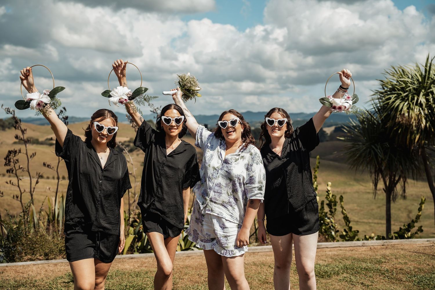 Four women in sunglasses raise white doves outdoors at a scenic countryside wedding celebration.