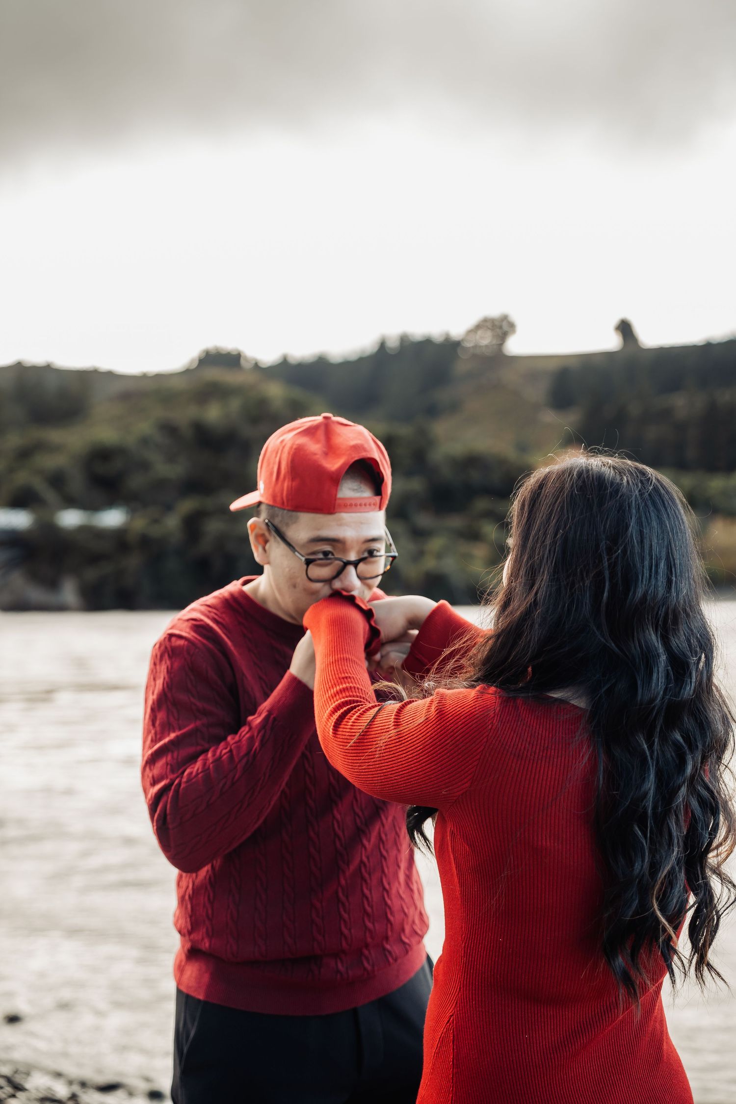 Man in red sweater and cap kissing woman's hand near a scenic waterfront.