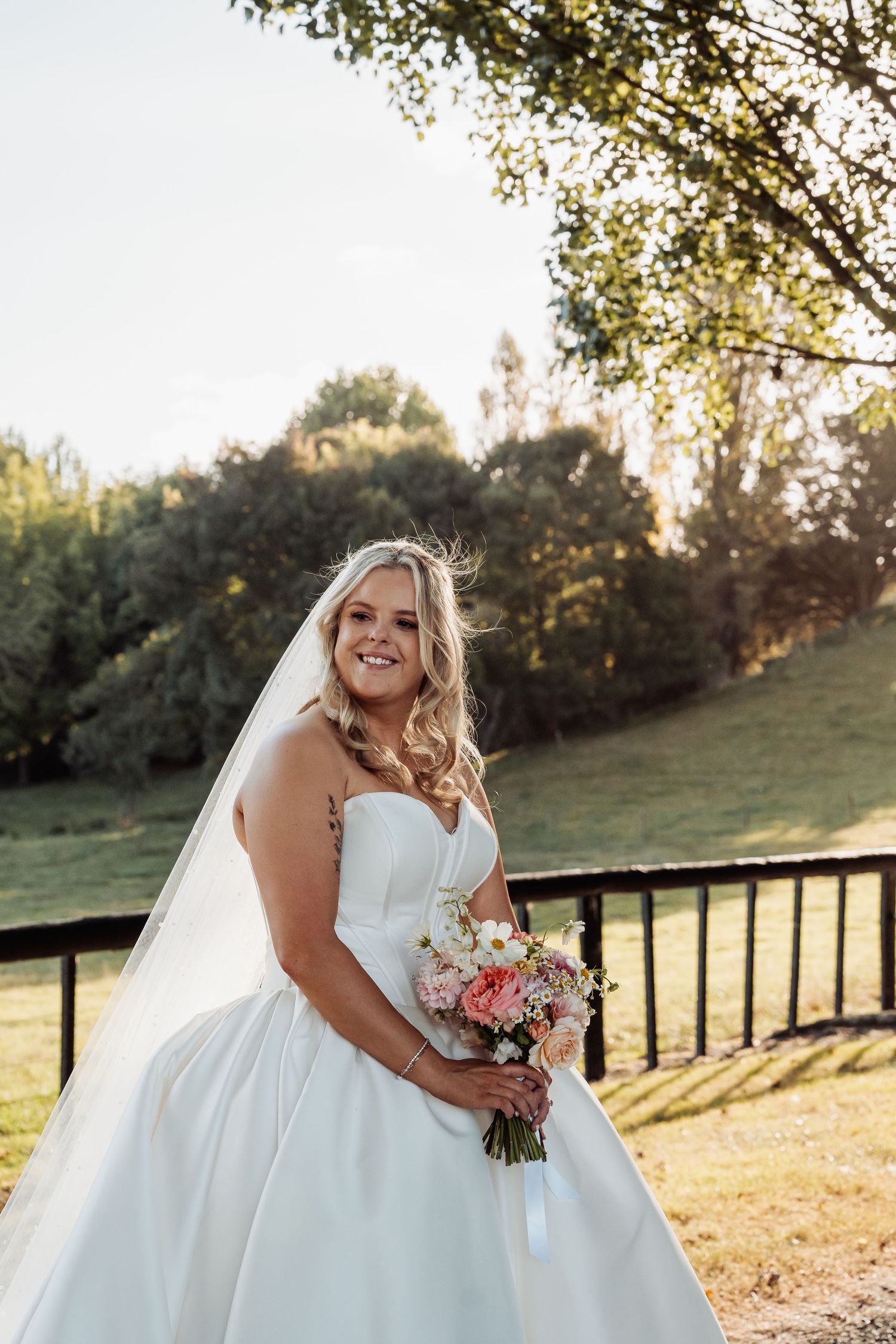 Radiant bride in strapless white gown holds pastel bouquet in sunny outdoor garden setting.
