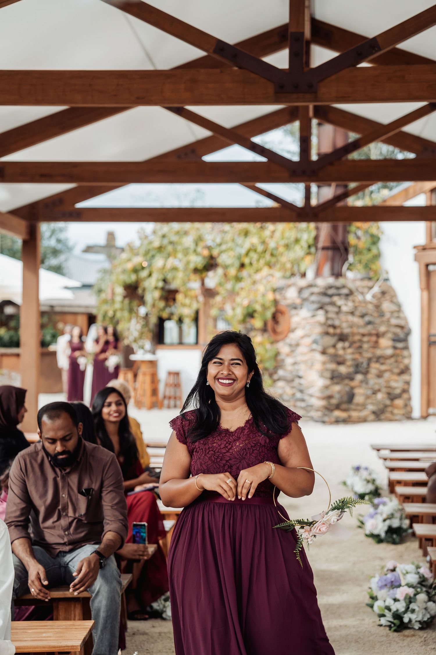 Smiling bridesmaid in burgundy lace dress walking down outdoor wedding aisle under wooden pergola.