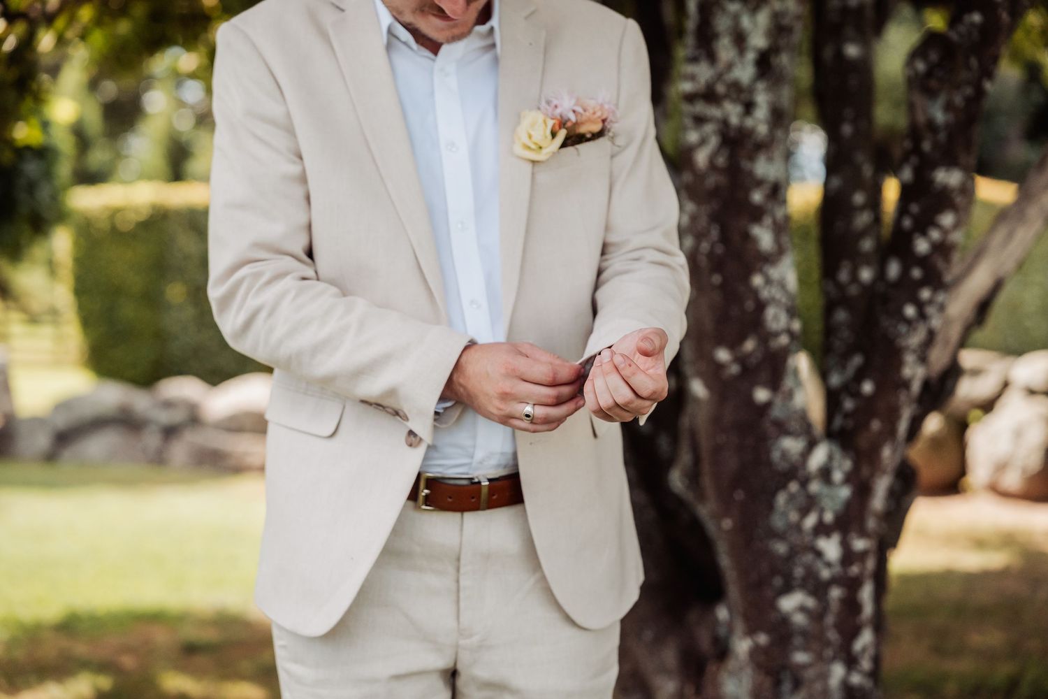 Groom in beige suit adjusting cufflinks with a floral boutonniere outdoors.