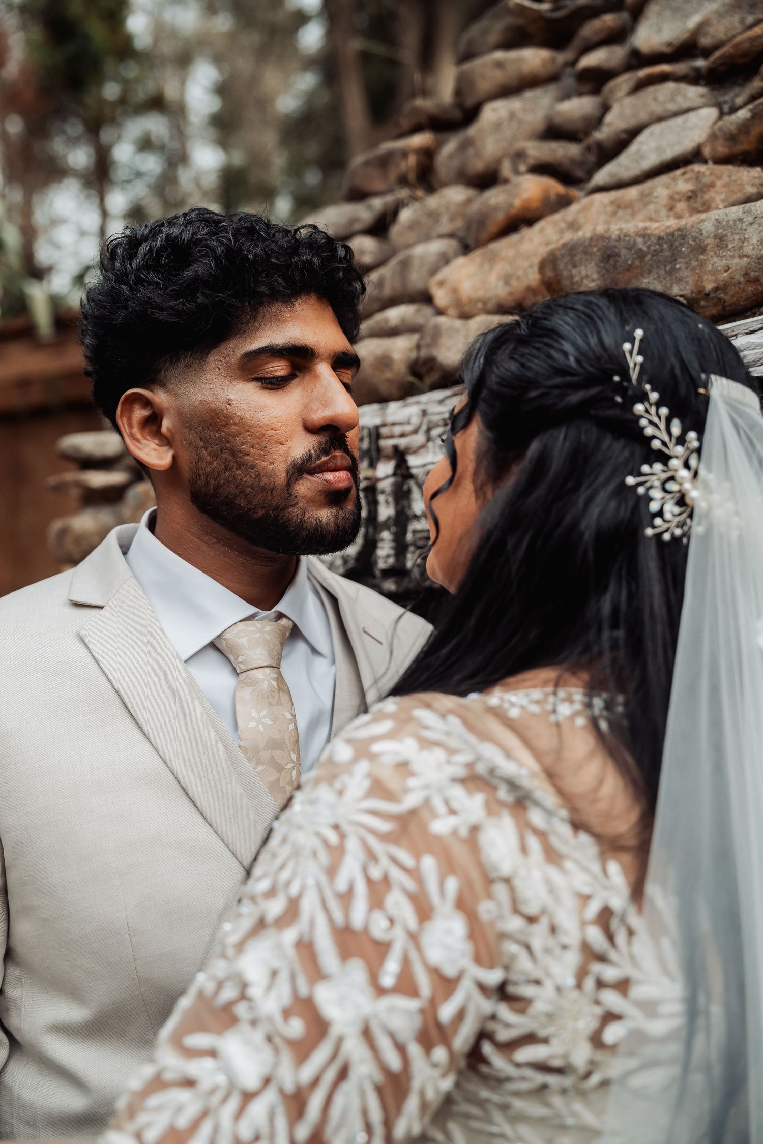 Groom in a beige suit leaning in close to bride in lace wedding dress near a stone wall.