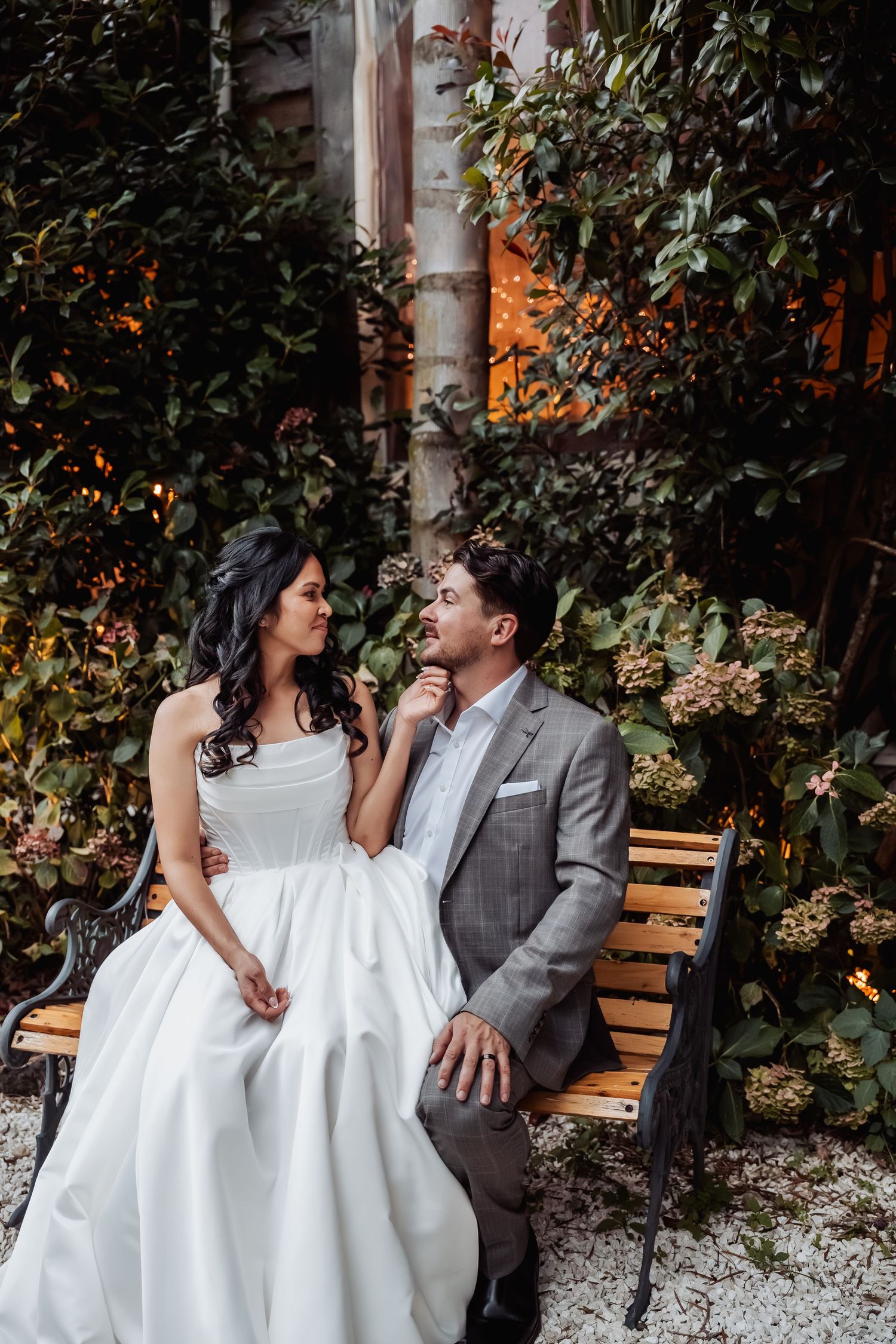 Bride and groom sit intimately on garden bench surrounded by lush greenery at twilight wedding reception.