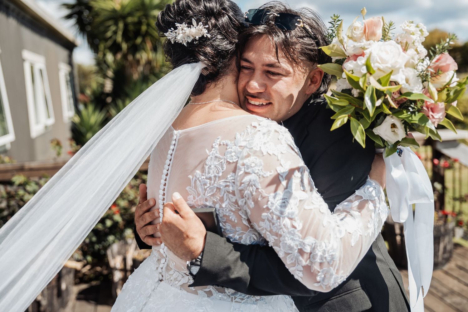 A joyful wedding embrace captured outdoors, with a flowing white lace gown and beautiful pink and white bouquet.
