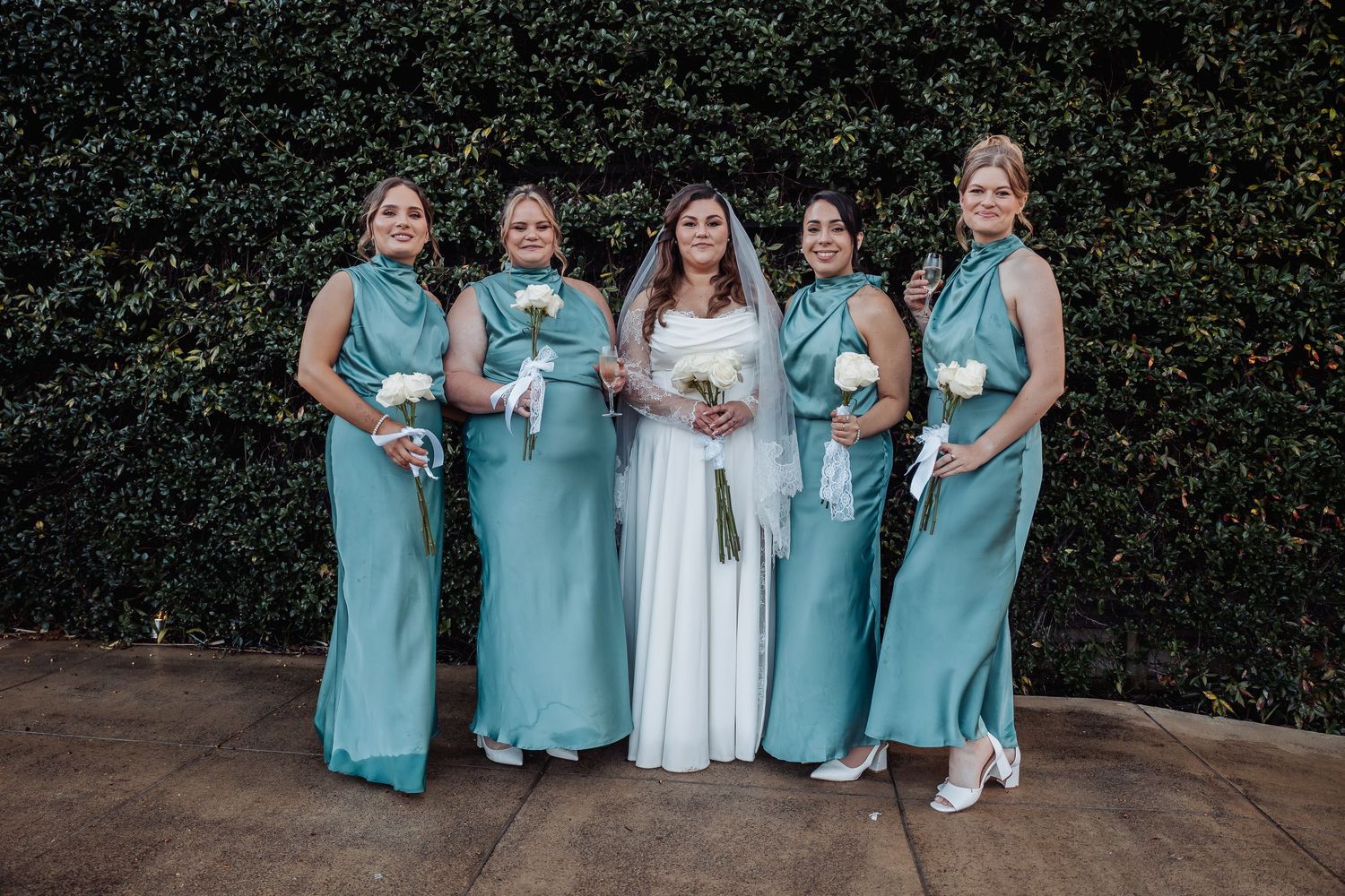 Wedding party in coordinated turquoise dresses poses together in front of lush green hedge backdrop.