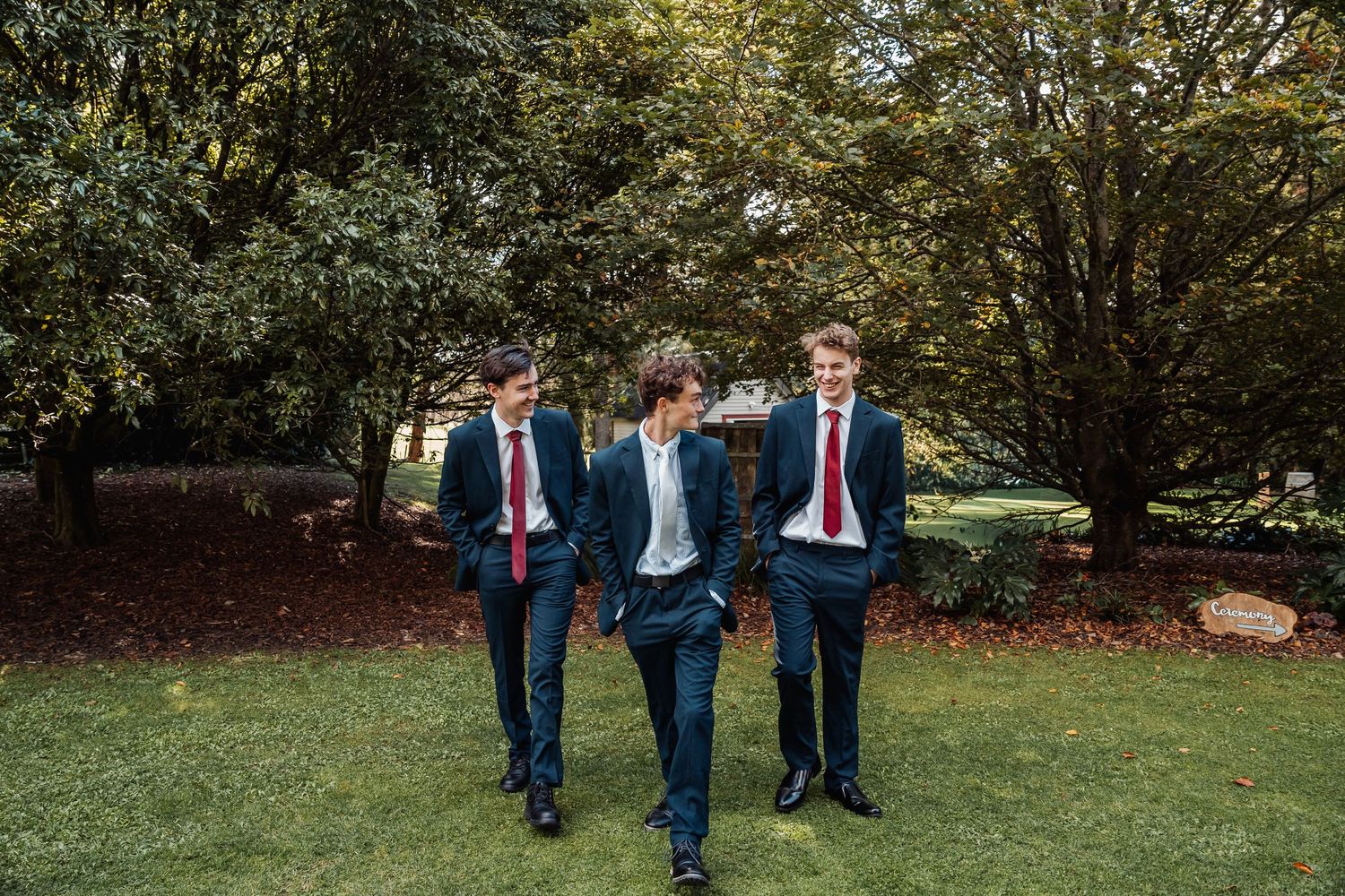 Three groomsmen in navy suits with red ties walk together across green lawn at wedding.