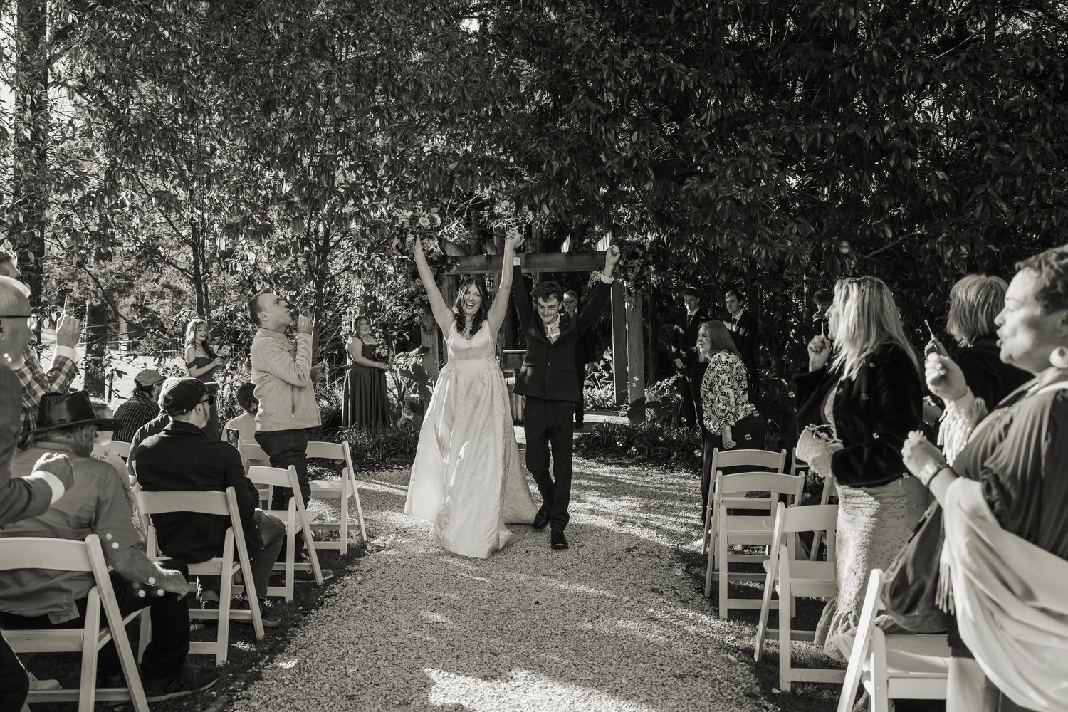 Black and white photo of jubilant newlyweds raising hands walking back down wedding aisle.