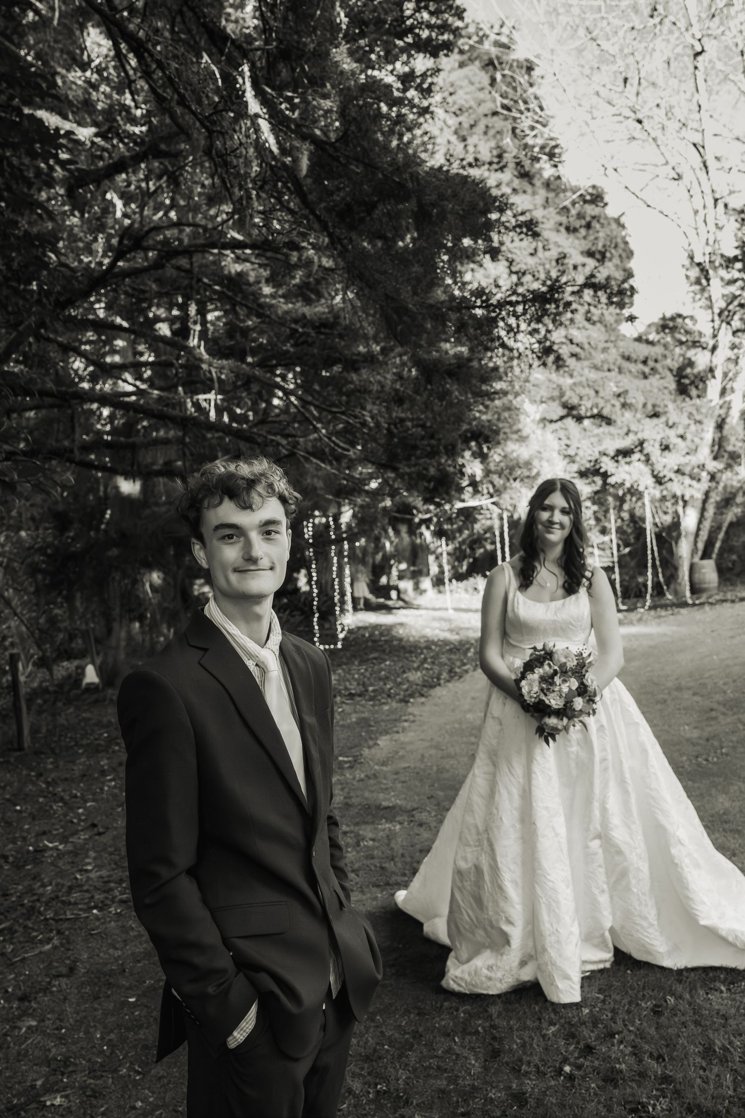Black and white portrait of groom in dark suit with bride holding bouquet on a tree-lined path.