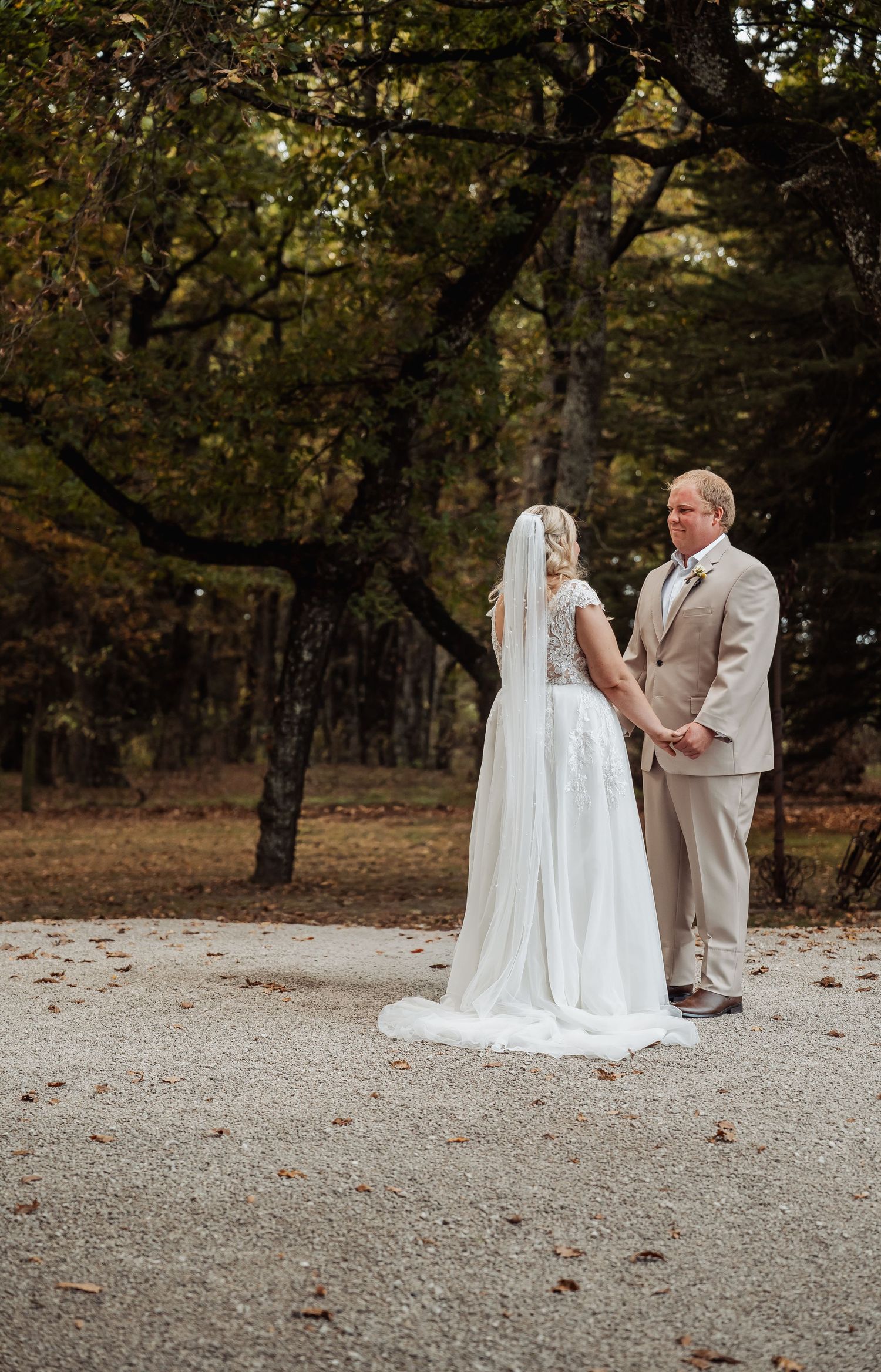 Bride and groom hold hands in a wooded autumn setting, bride wearing a long white gown with veil.