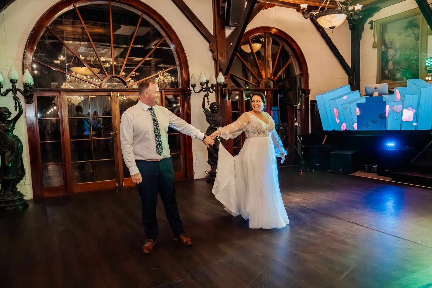 Wedding guests dance and celebrate on a dark wooden dance floor under rustic wooden beams with twinkling lights.