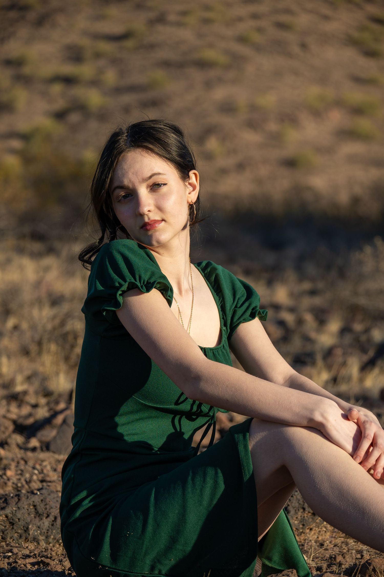 A lady in a green dress is sitting down in the middle of the desert and smiling.