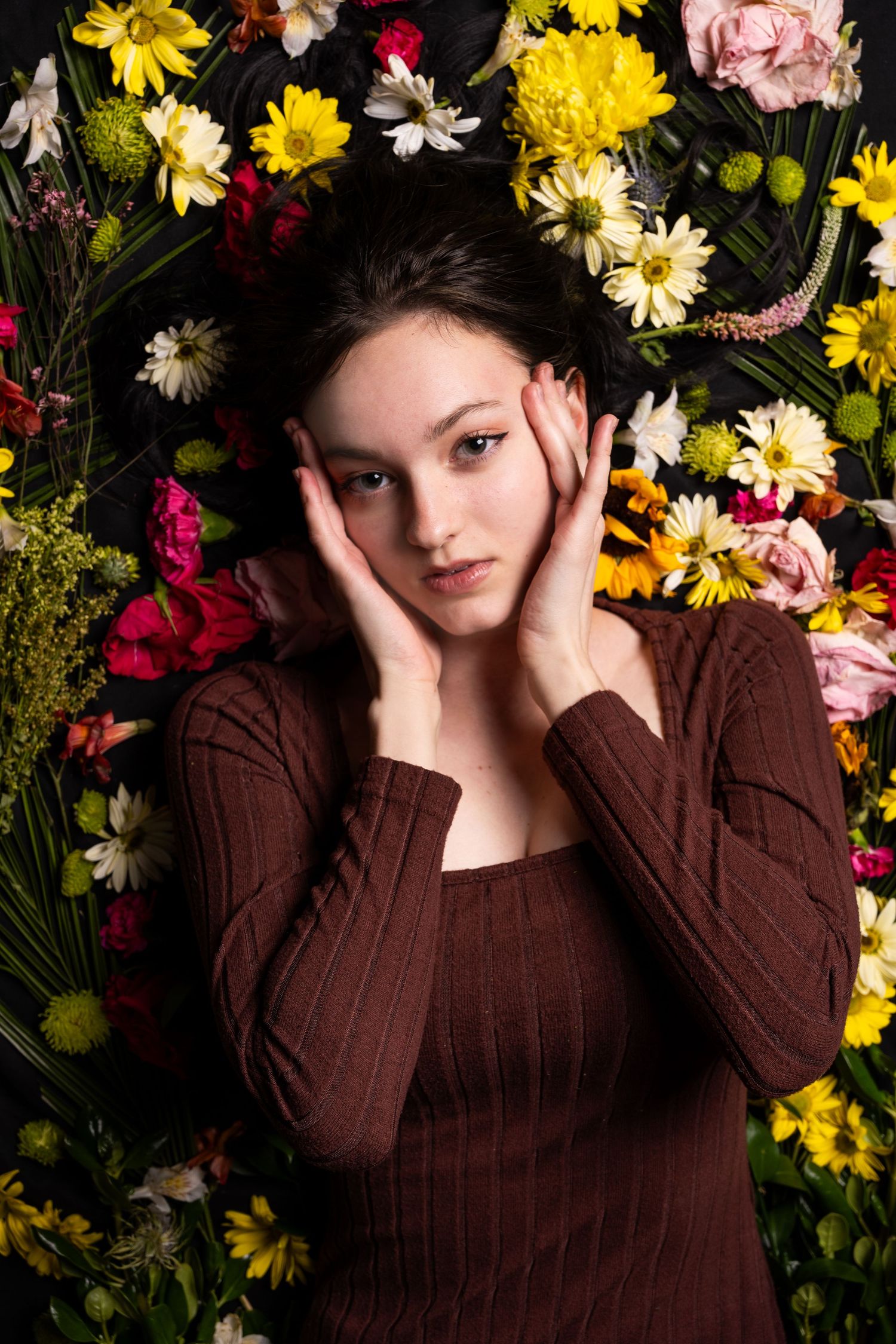 A lady is laying down with flowers around her. She has her hands on her face and framing her face. She has a serious look on her face.