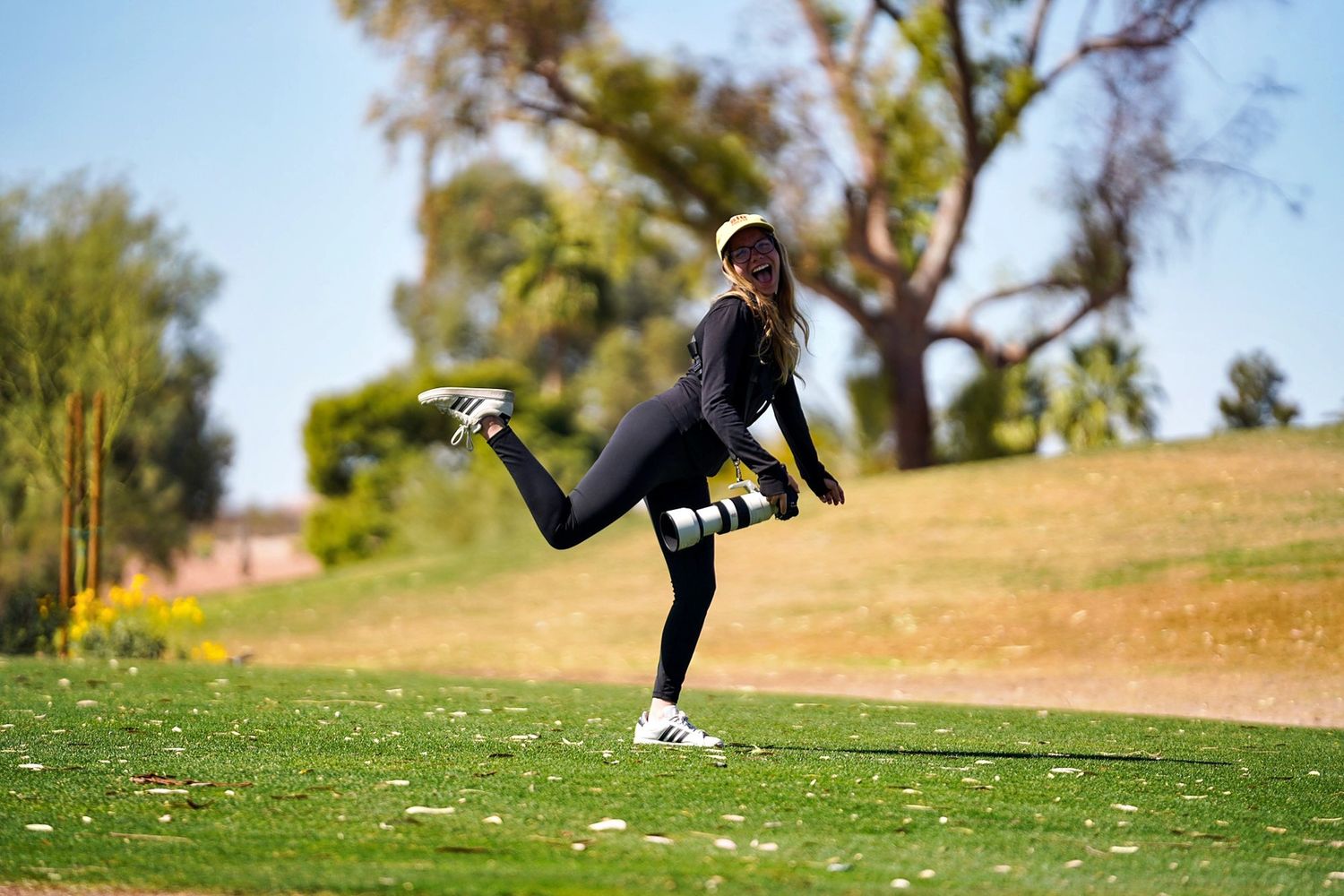 Danielle Gantt holding a camera and standing on one leg smiling.