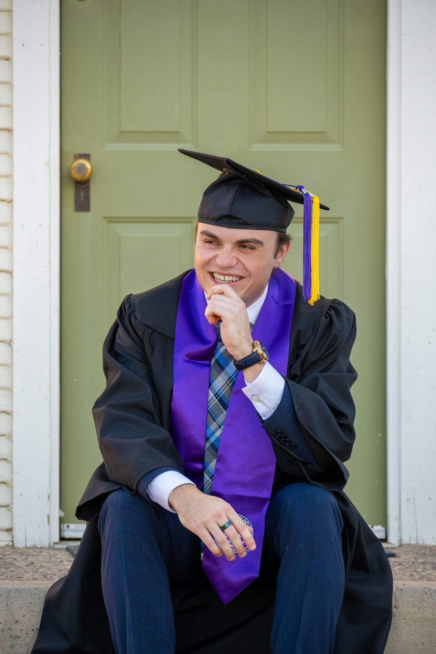 Man in graduation attire from Grand Canyon University. He is sitting while looking off into the distance and laughing.