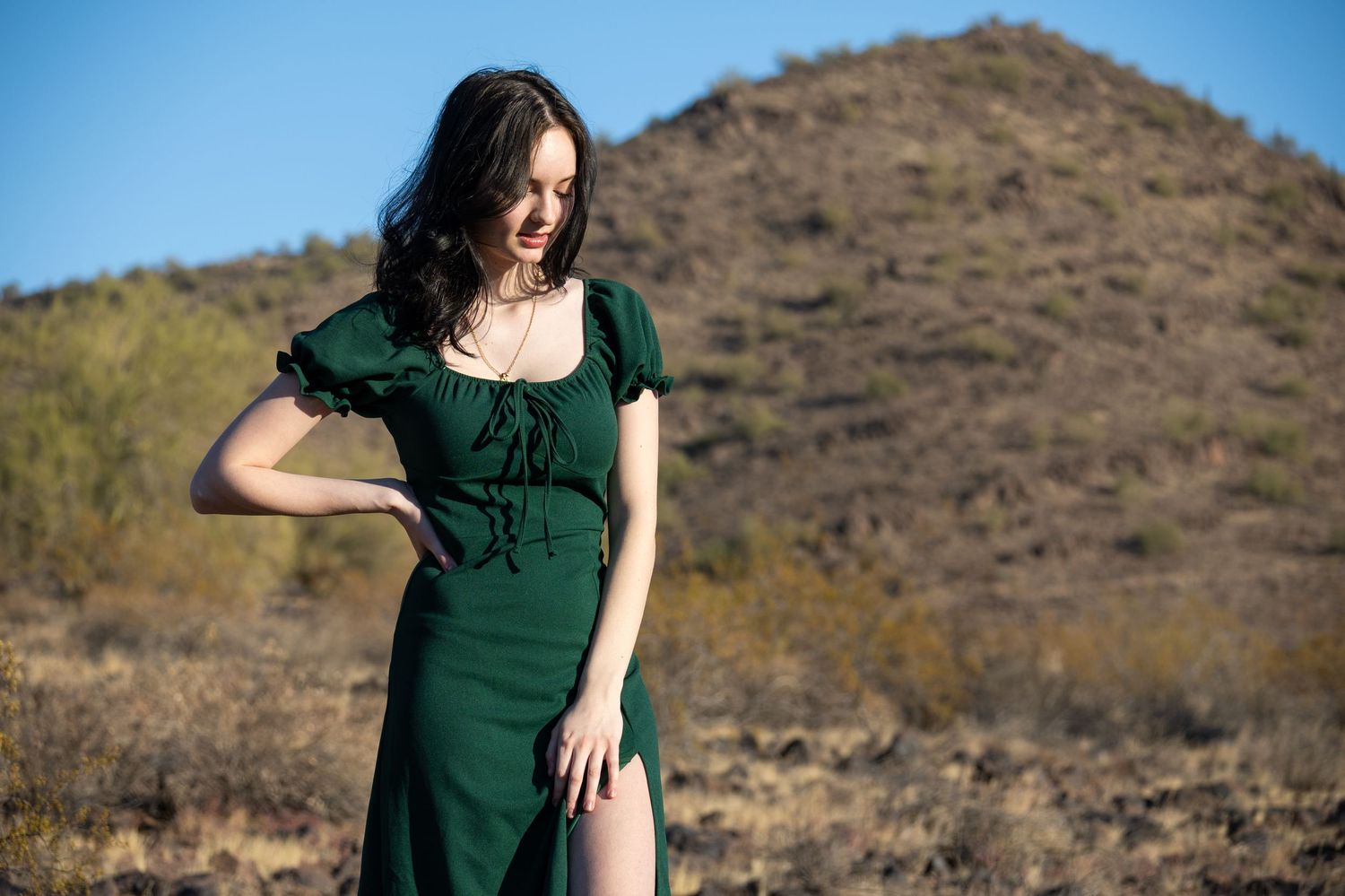 A lady in a green dress is standing in the desert. She has one hand on her hip and the other hangs while she looks down.