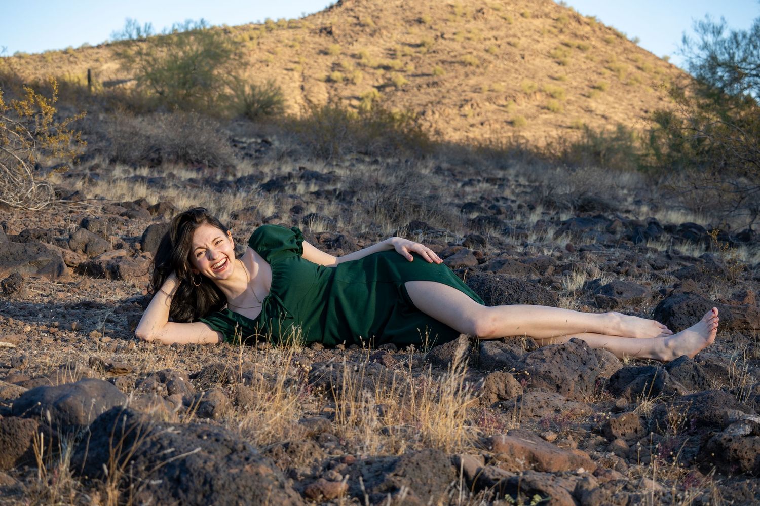 A lady in a green dress is laying down in the middle of the desert while laughing and smiling.
