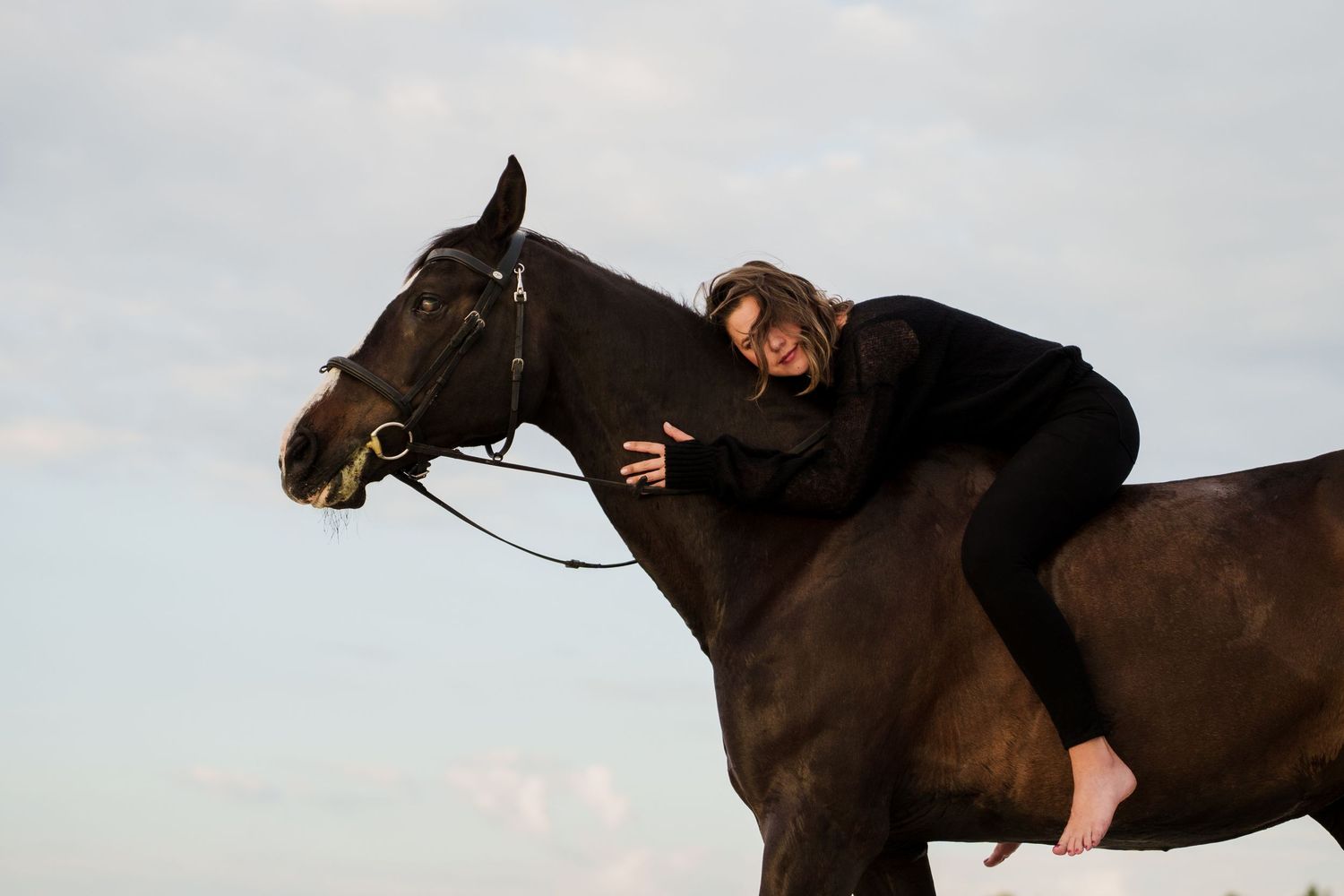 Woman in black outfit lying across a dark brown horse against a cloudy sky.