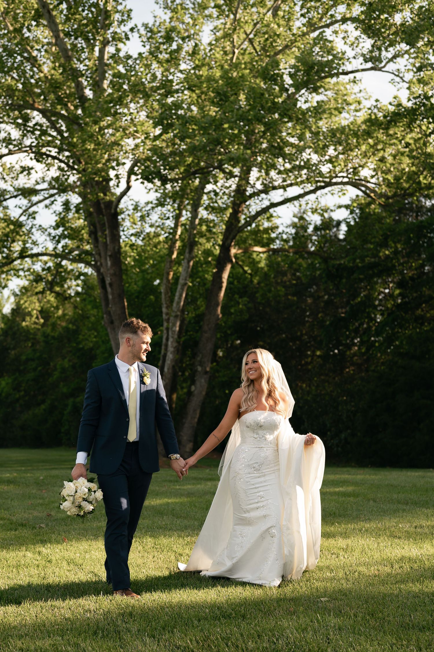 Newlywed couple poses together in a dreamy sunset field with tall trees creating a romantic outdoor setting.