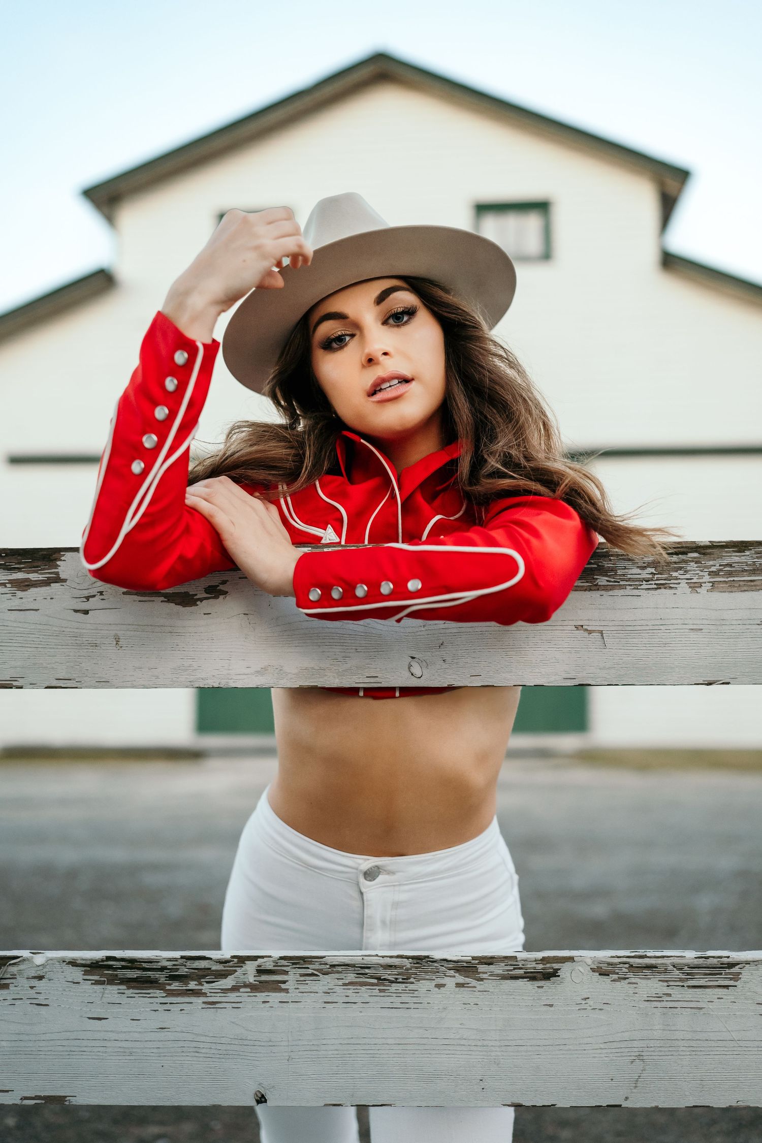 Fashion portrait featuring a red cropped jacket, white pants, and western hat against a white fence.