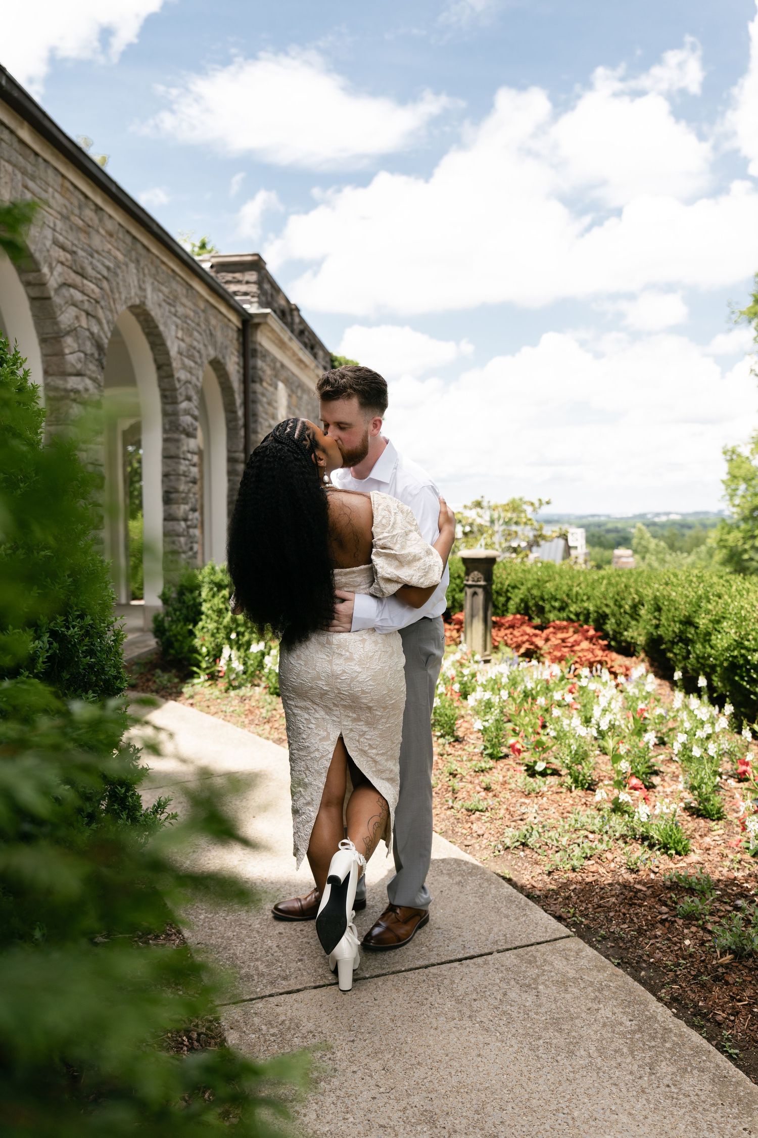 Couple sharing a romantic kiss on a garden pathway beside a stone building, surrounded by colorful flowers on a sunny day.