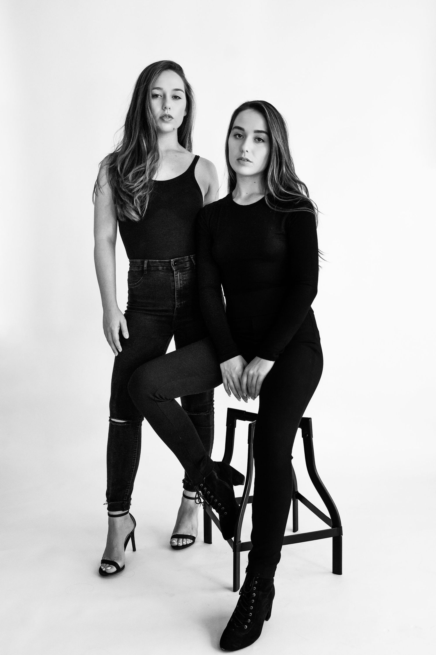 Two women in black outfits pose together in minimalist studio setting.